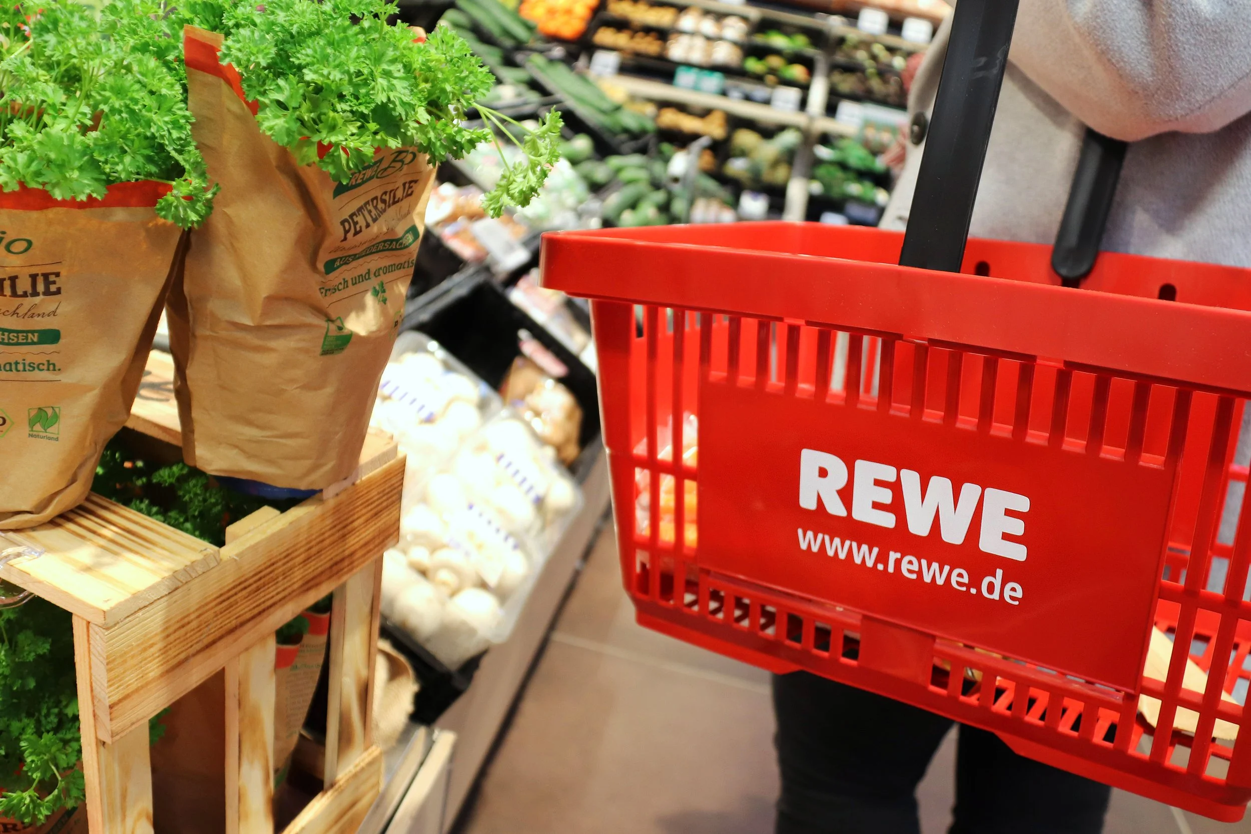 Einkaufskorb mit der Aufschrift 'REWE' neben Obst- und Gemüseständen im Supermarkt, mit einer Hand, die den Korb hält.