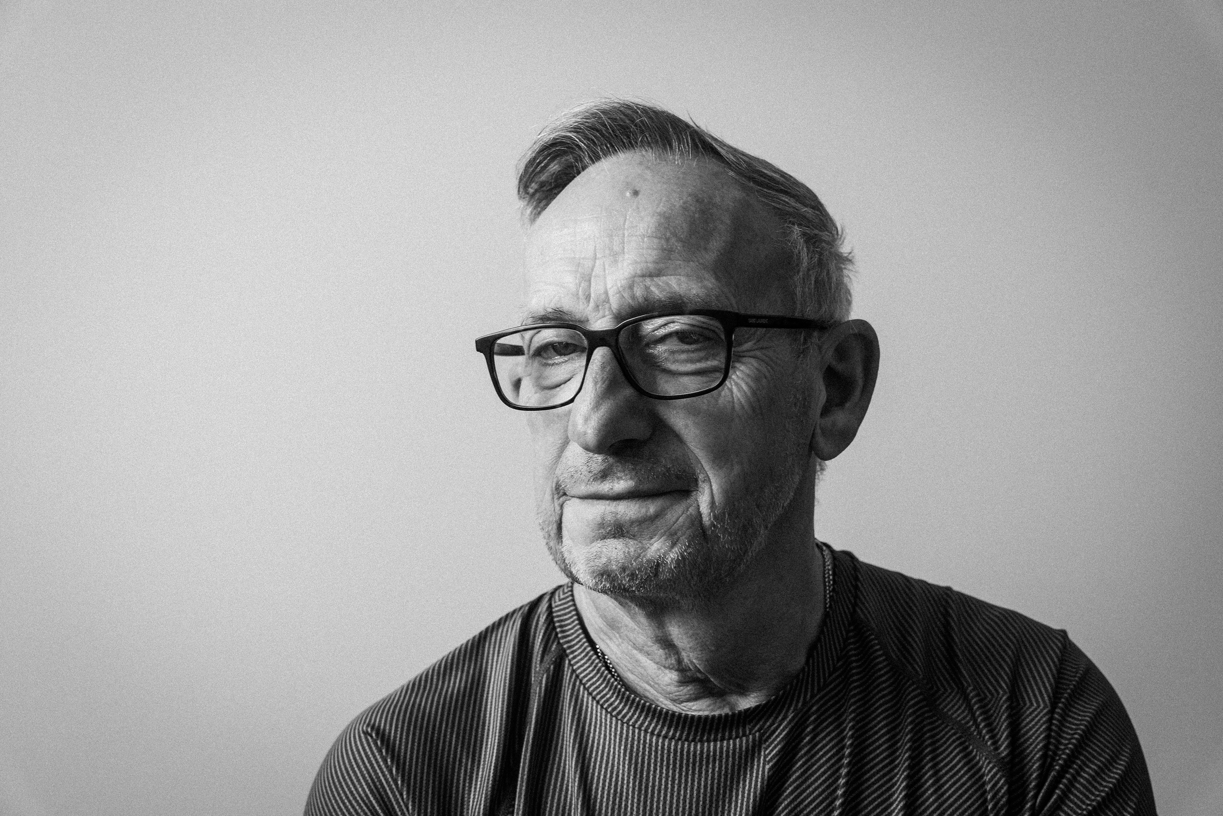 Schwarz-weiß Foto eines älteren Mannes mit kurzen, grauen Haaren, Brille und einem freundlichen Gesichtsausdruck, trägt ein gestreiftes Hemd.
