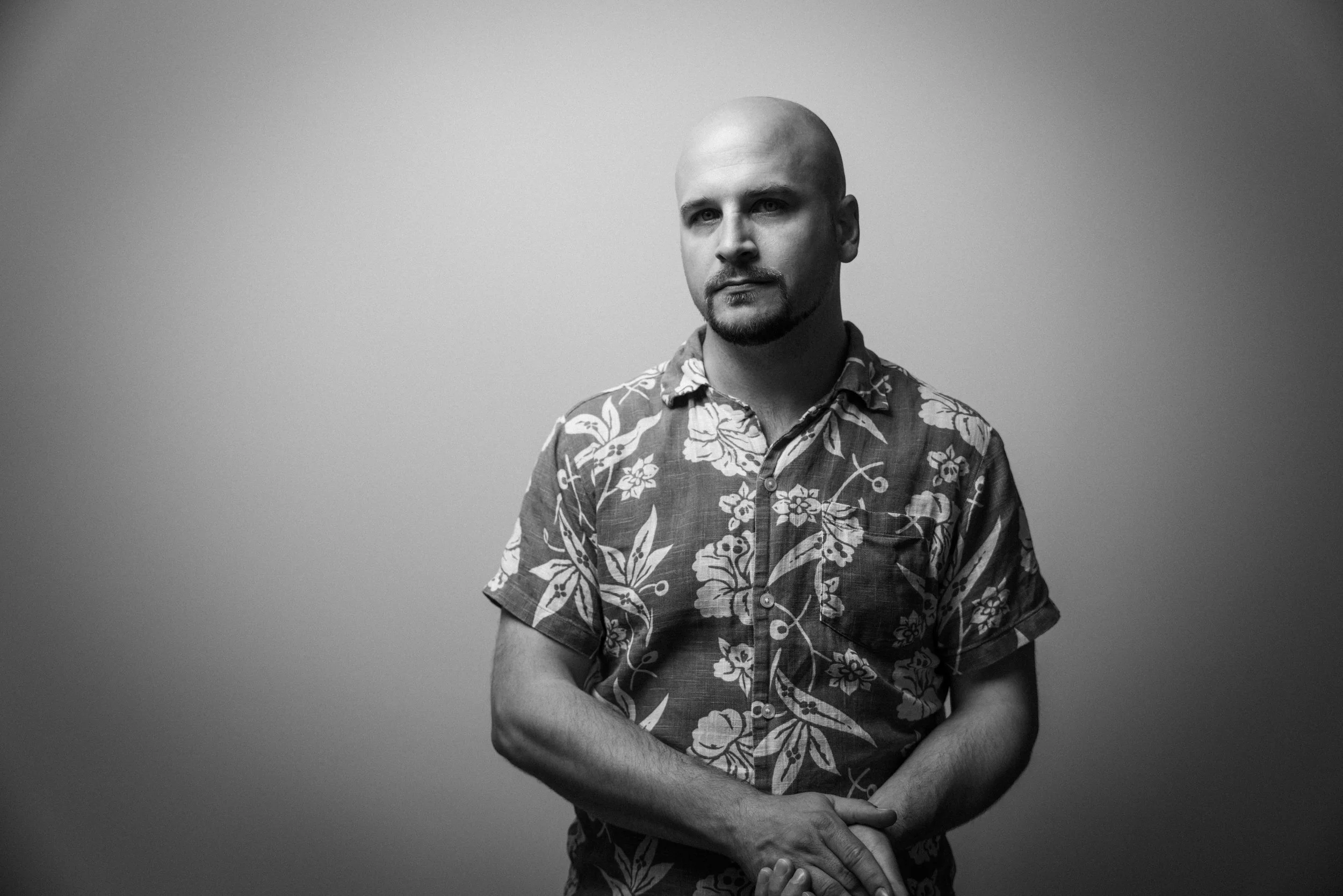 Schwarz-Weiß-Foto eines Mannes mit Glatze und Bart, der ein Hawaihemd trägt, vor grauem Hintergrund.