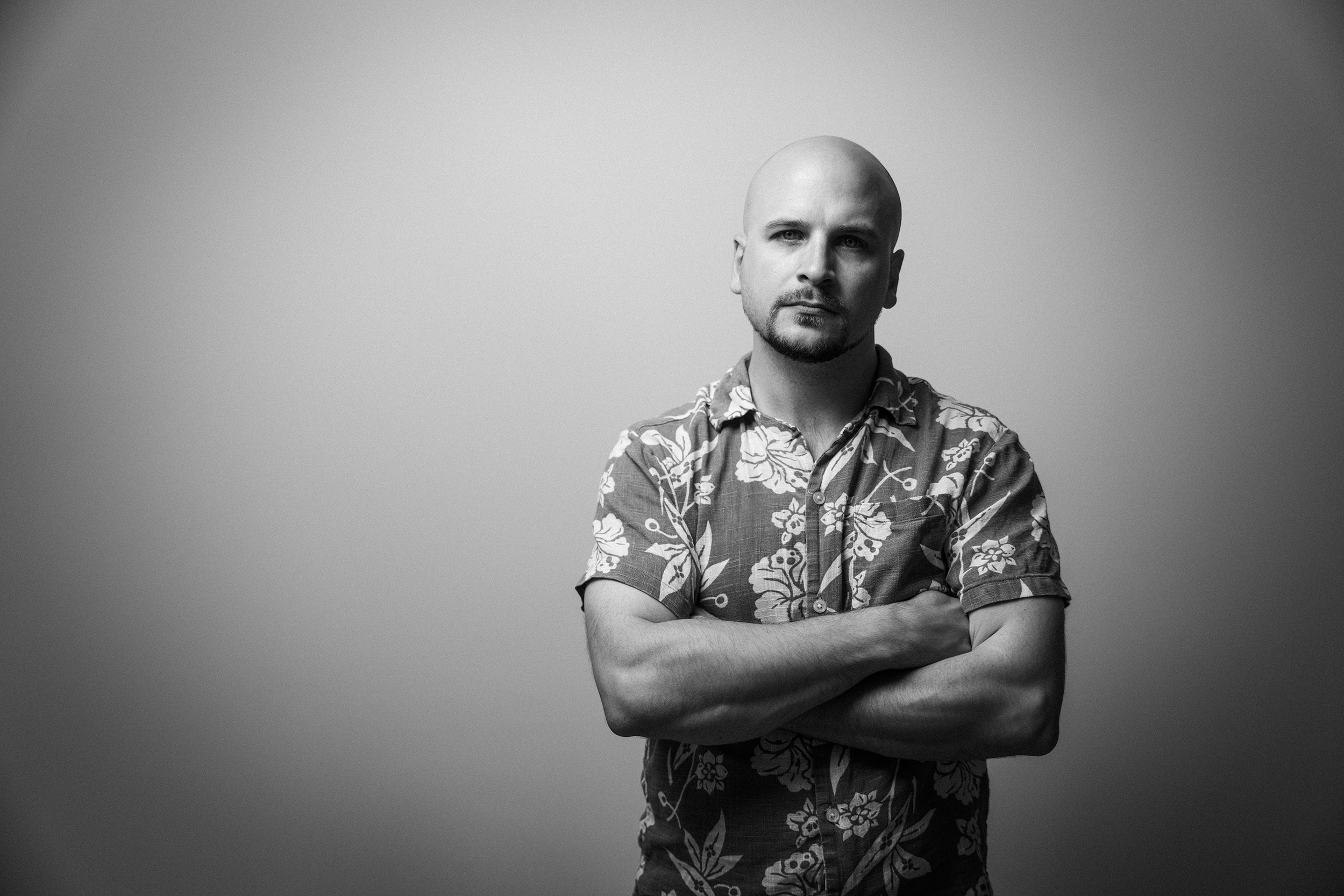 Schwarz-Weiß-Fotografie eines Mannes mit glattem Kopf, Schnurrbart und Bart, der ein Aloha-Hawaii-Hemd trägt, die Arme verschränkt, vor einem neutralen grauen Hintergrund.