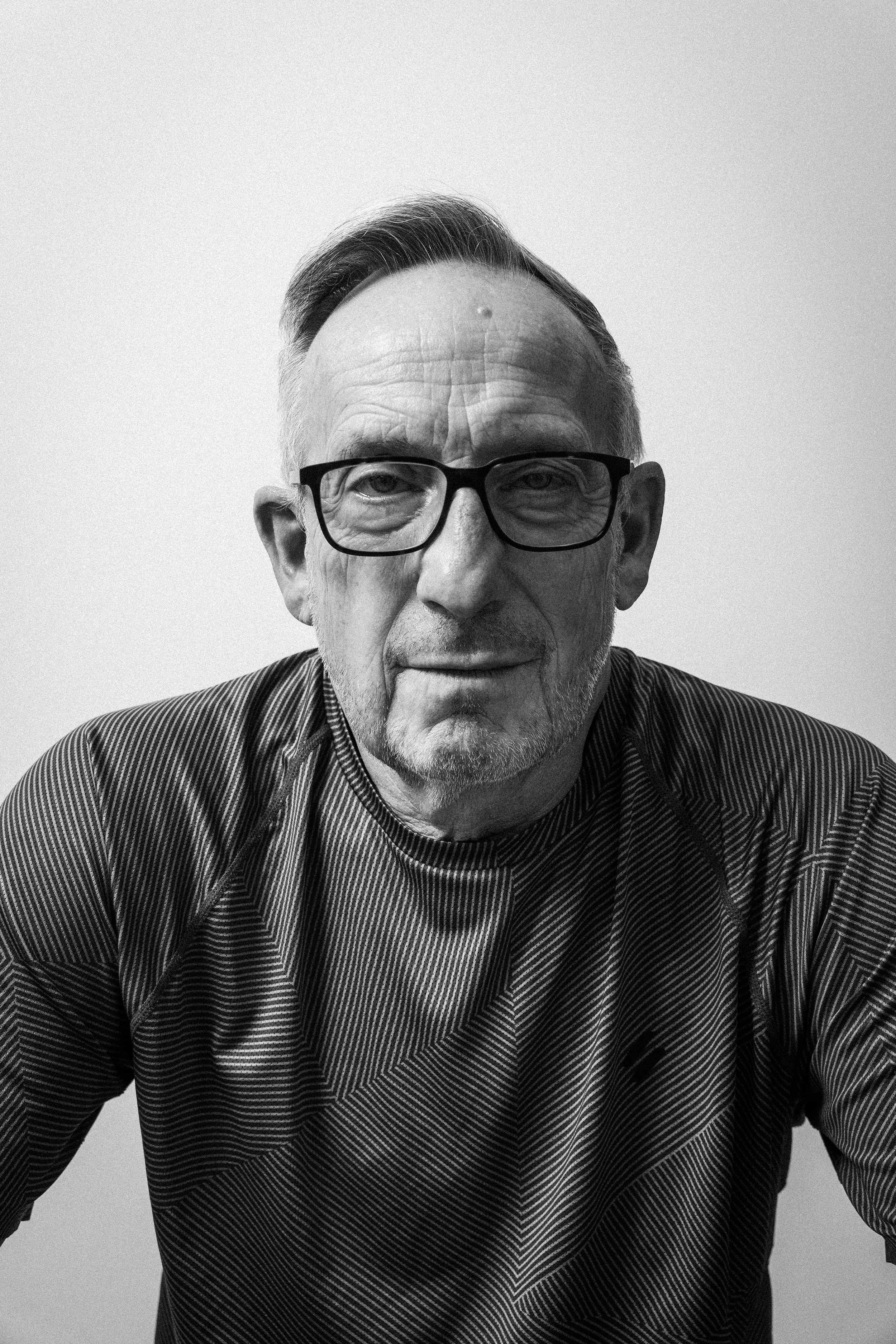 Schwarz-Weiß-Fotografie eines älteren Mannes mit Brille, lächelnd, bekleidet mit einem gestreiften Hemd.