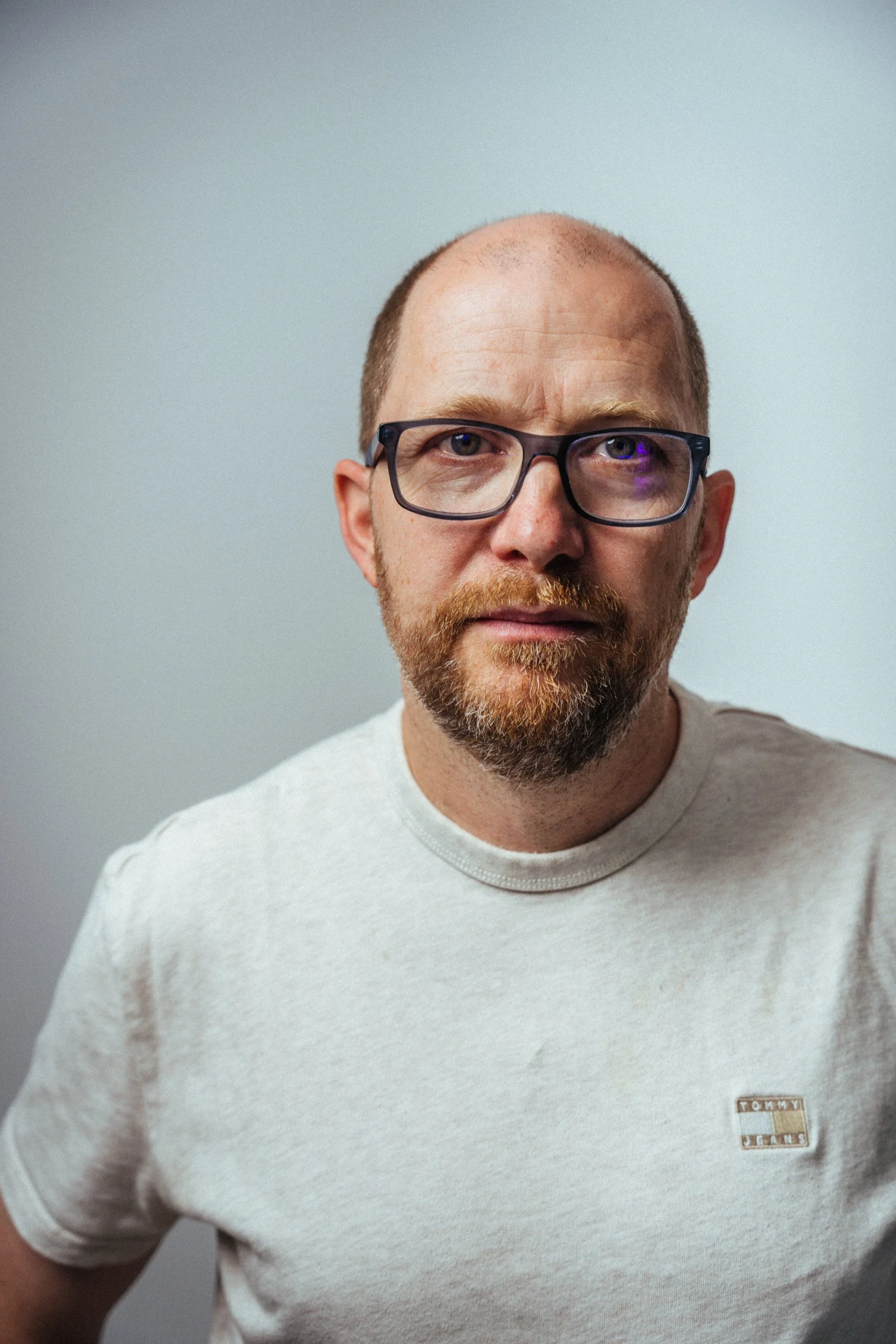 Porträt eines Mannes mit kurzgeschnittenem Haar, Brille, Bart und einem weißen T-Shirt, vor einem grauen Hintergrund.