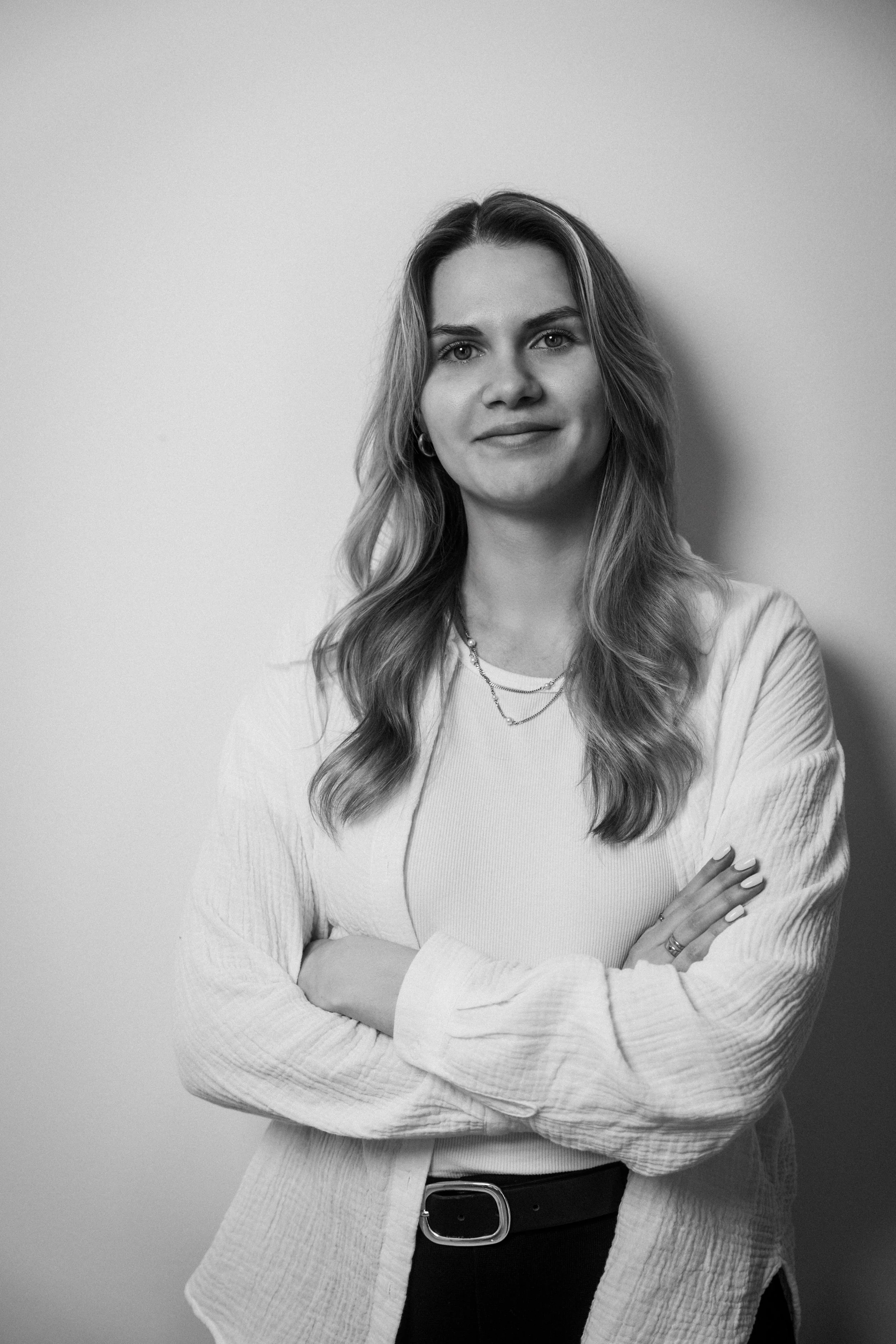 Schwarz-weiße Portraitaufnahme einer jungen Frau mit langen, gewellten Haaren, die lächelt und die Arme verschränkt trägt eine minimalistische Halskette, einen Gürtel und ein locker sitzendes Oberteil. Jenna Sauer in  Das Wilde Weib – Das Musical · B