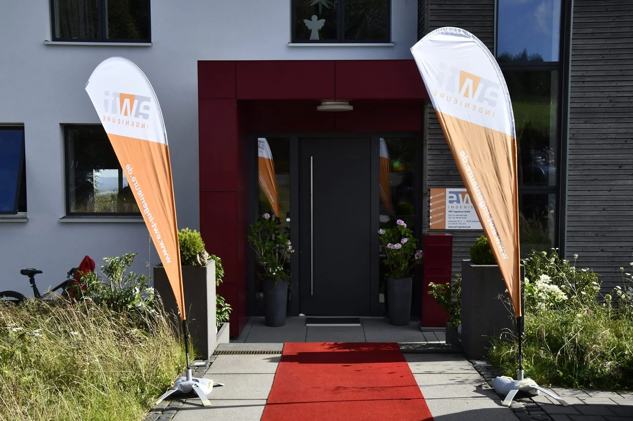 Der Eingang eines modernen Gebäudes mit roten Akzenten und zwei orangenen Fahnen, die Werbung für einen Ingenieurbüro zeigen, sowie einem roten Läufer auf dem Gehweg vor der Tür, umgeben von Pflanzen und Blumen.
