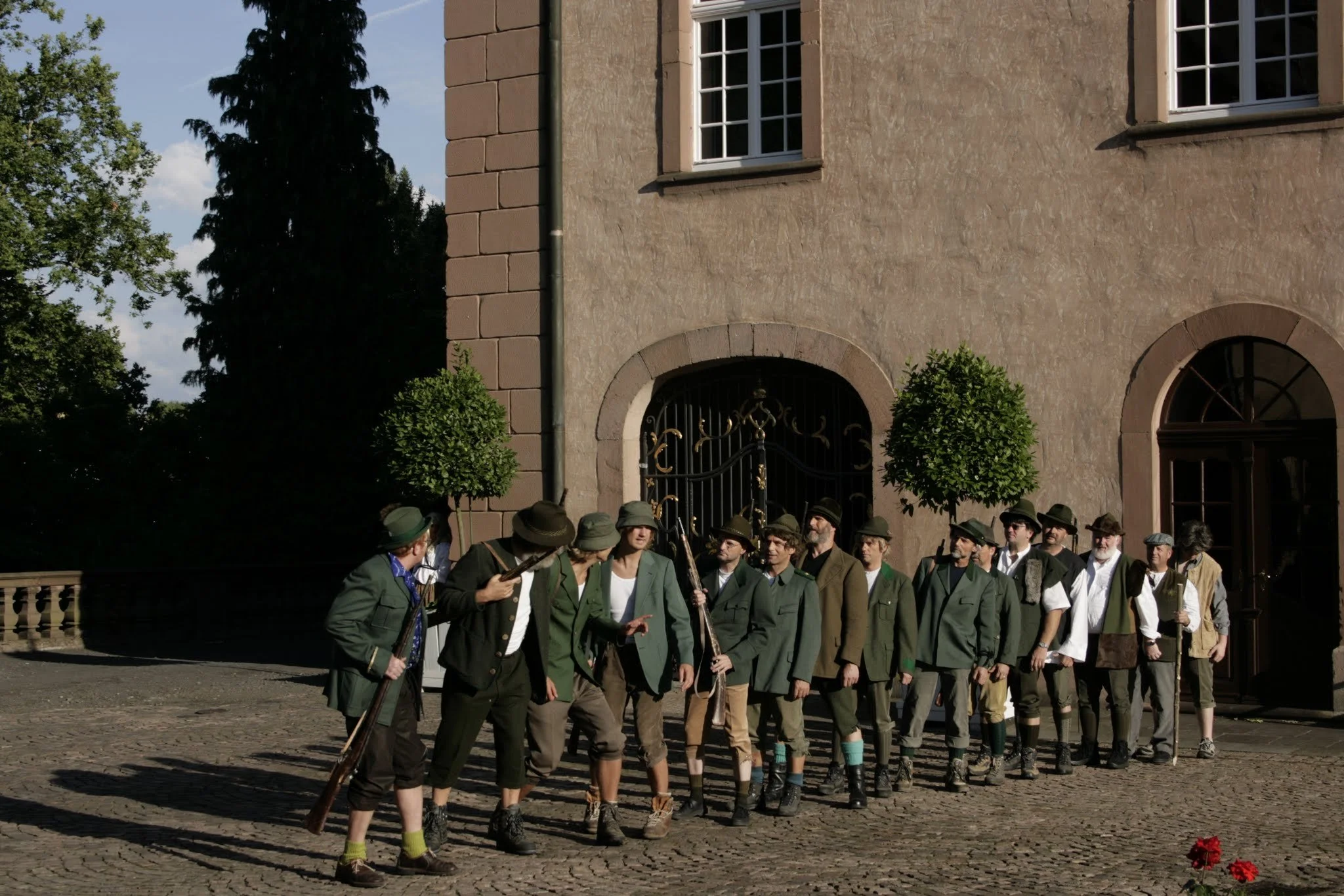 Eine Gruppe von Menschen, die in historischen Kostümen im Freien stehen, vermutlich bei einem Theater- oder Kulturfestival. Sie tragen grüne und braune Kleidung mit Hüten und Stöcken, stehen in einer Reihe vor einem alten Gebäude mit Bäumen im Hinter