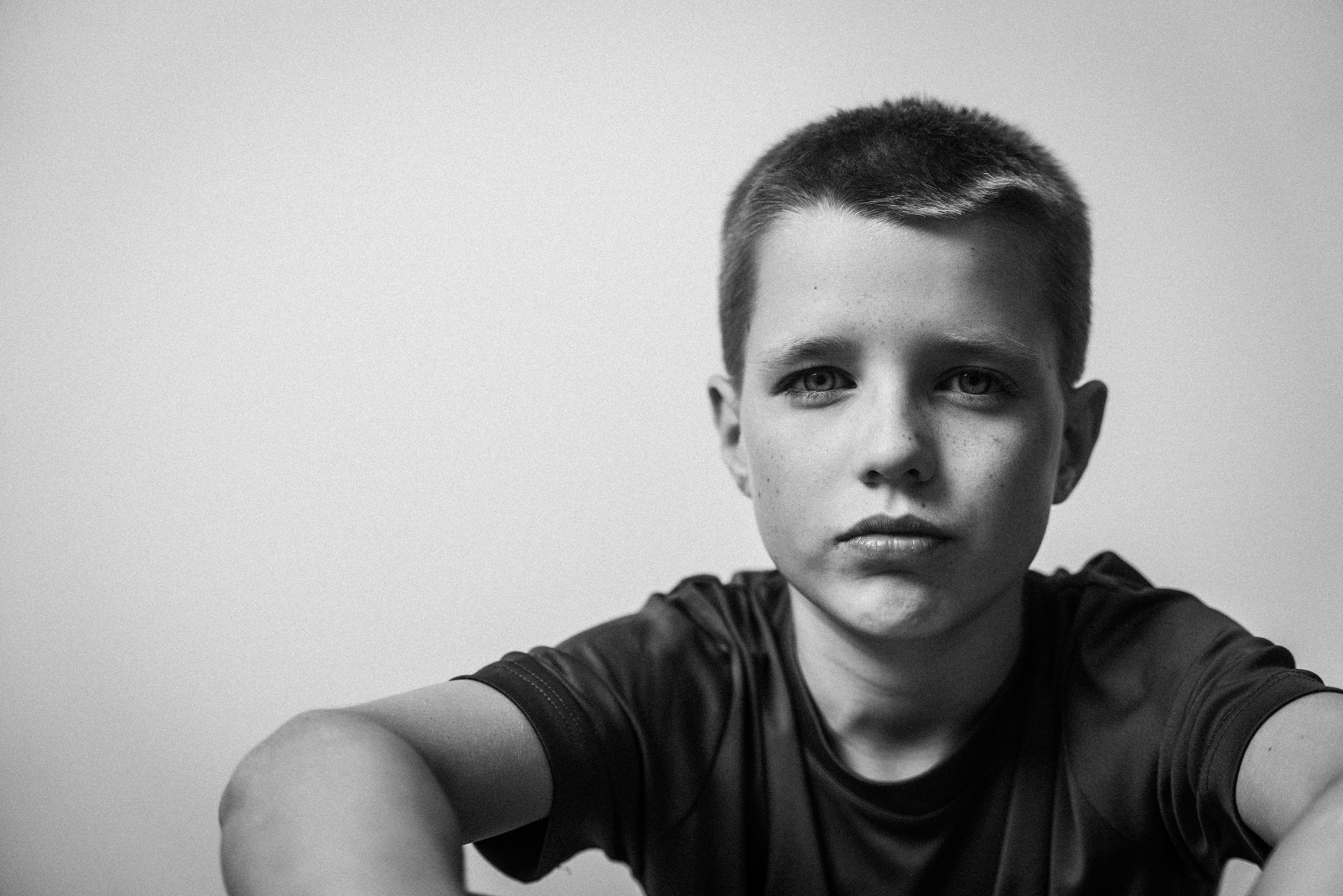 Schwarz-weißporträt eines Jungen mit kurzen dunklen Haaren, ernstem Ausdruck, schwarzem T-Shirt, vor hellem Hintergrund.