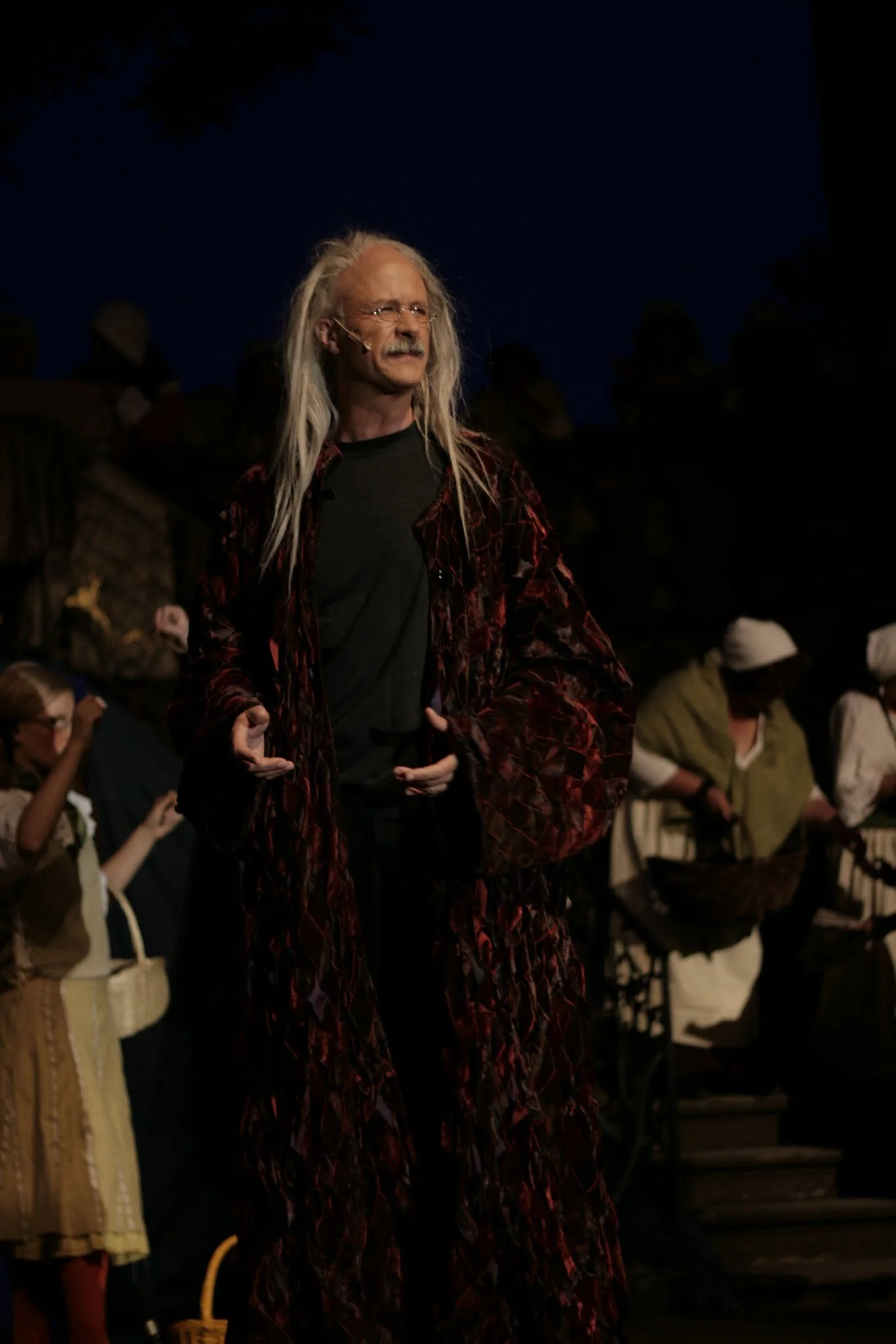 Mann mit langen grauen Haaren, dunklem Hemd und rotem, gewebtem Umhang bei einer Theateraufführung oder Feier im Freien bei Nacht.