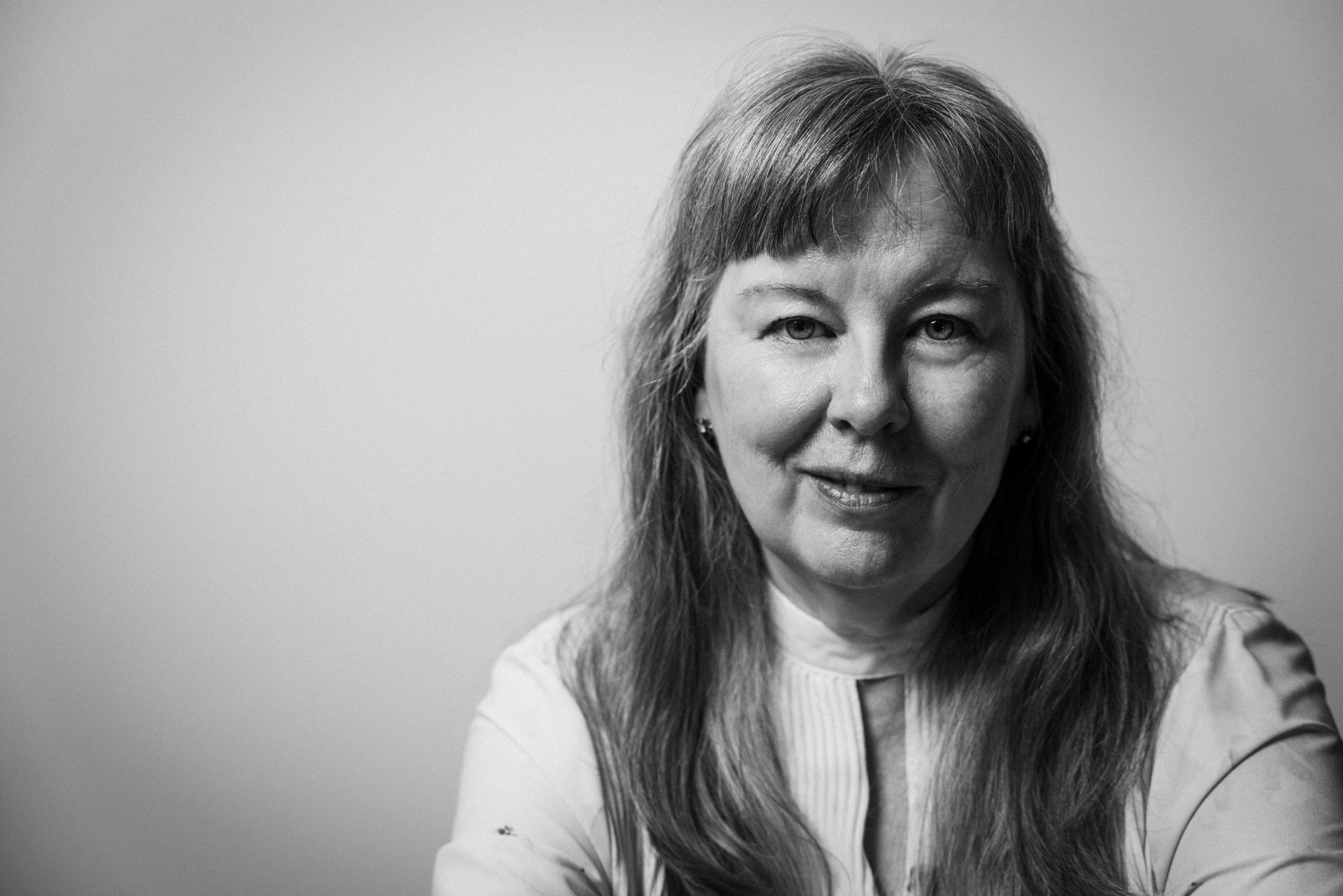 Schwarz-Weiß-Fotografie einer lächelnden Frau mit langen Haaren, die einen weißen Pullover trägt, vor einem neutralgrauen Hintergrund.