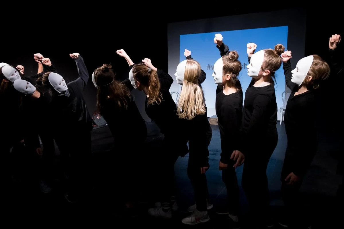 Eine Gruppe von Menschen trägt weiße Masken und schwarze Kleidung, sie stehen in einer Reihe auf einer Bühne, einige heben die Fäuste in die Luft. Im Hintergrund ist ein Bildschirm zu sehen.