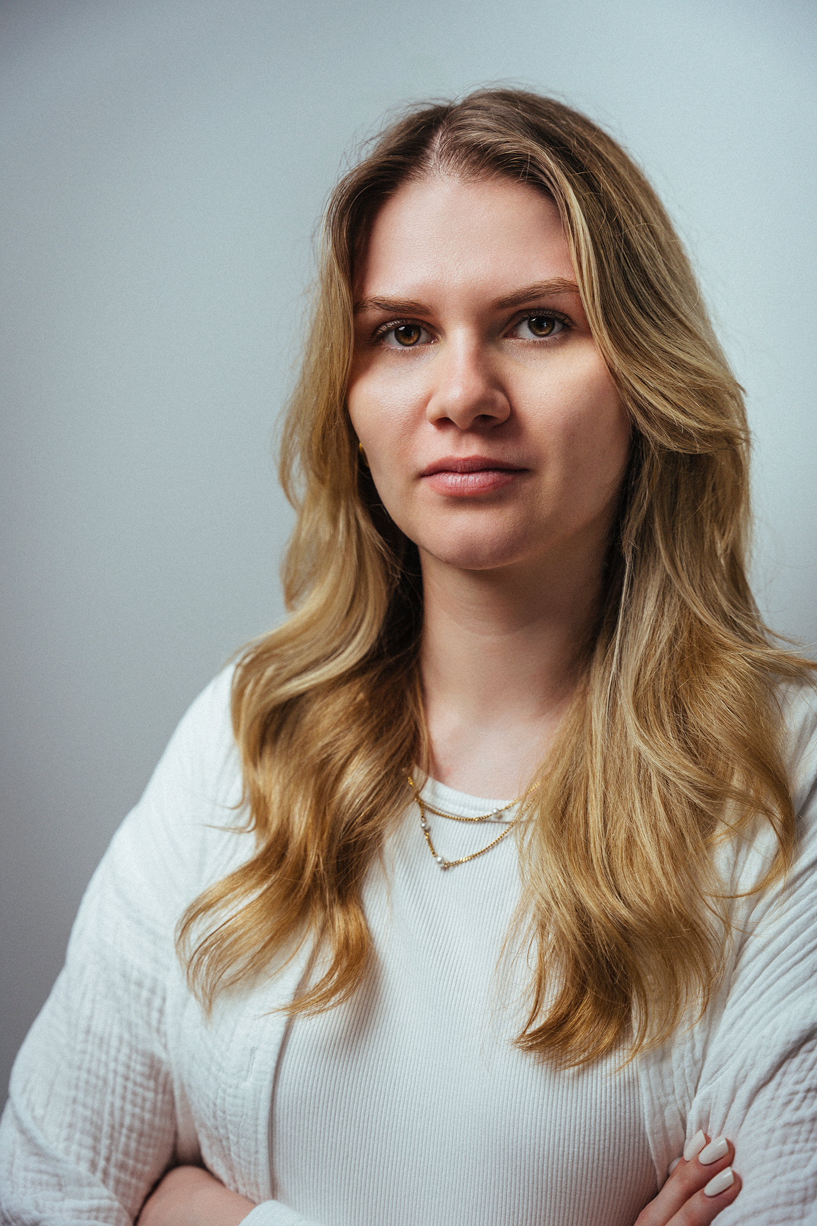 Ein junges Mädchen mit langem, gewelltem, hellblondem Haar, das einen weißen Pulli trägt und sich mit verschränkten Armen vor einem neutralen grauen Hintergrund befindet. Jenna Sauer  in  Das Wilde Weib – Das Musical · Birsteiner Festspiele 2027