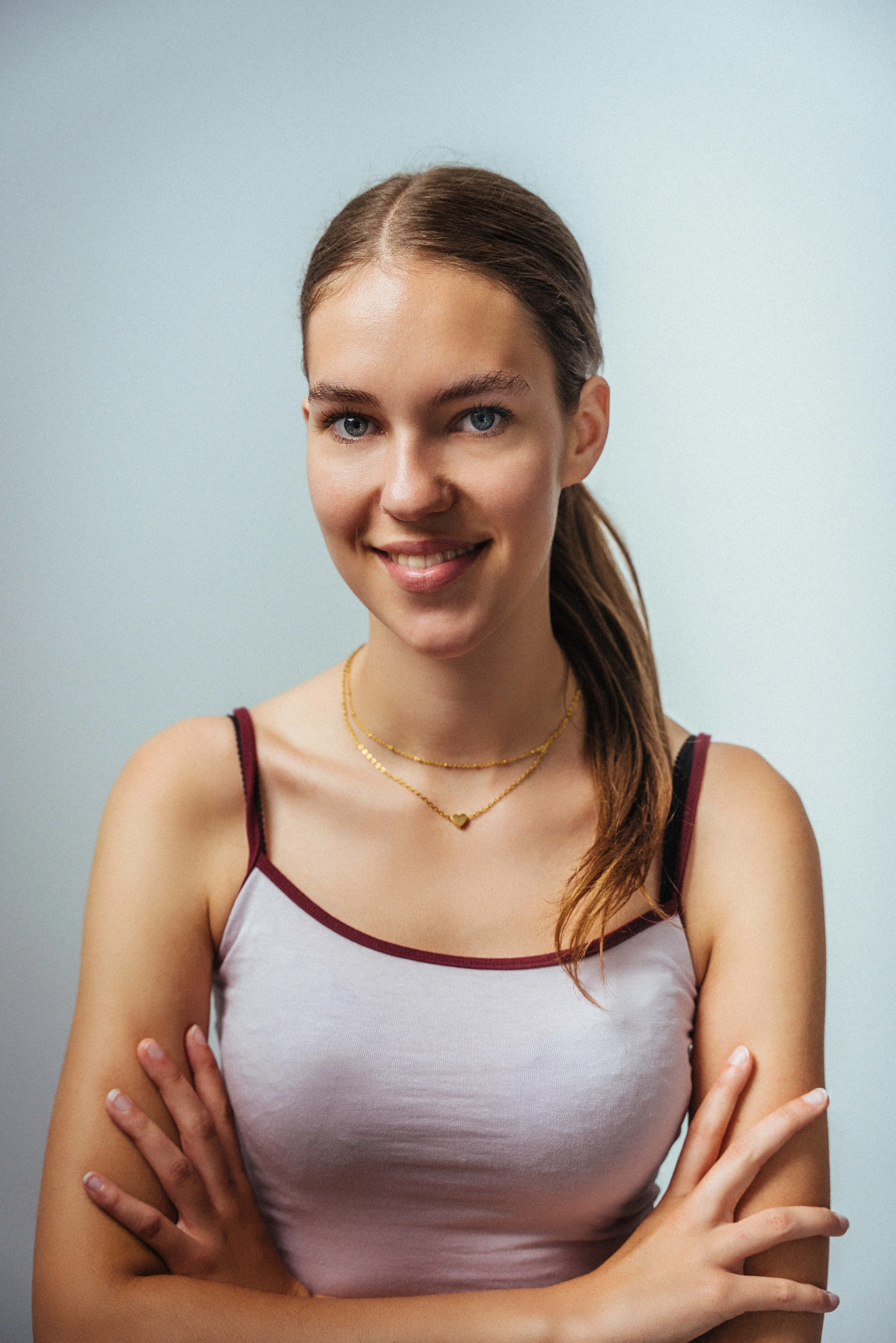 Junge Frau trägt eine weiße Top mit roten und schwarzen Akzenten, goldene Halsketten und hat braune Haare in einem lockeren Pferdeschwanz. Sie steht vor einem hellgrauen Hintergrund und lächelt leicht.