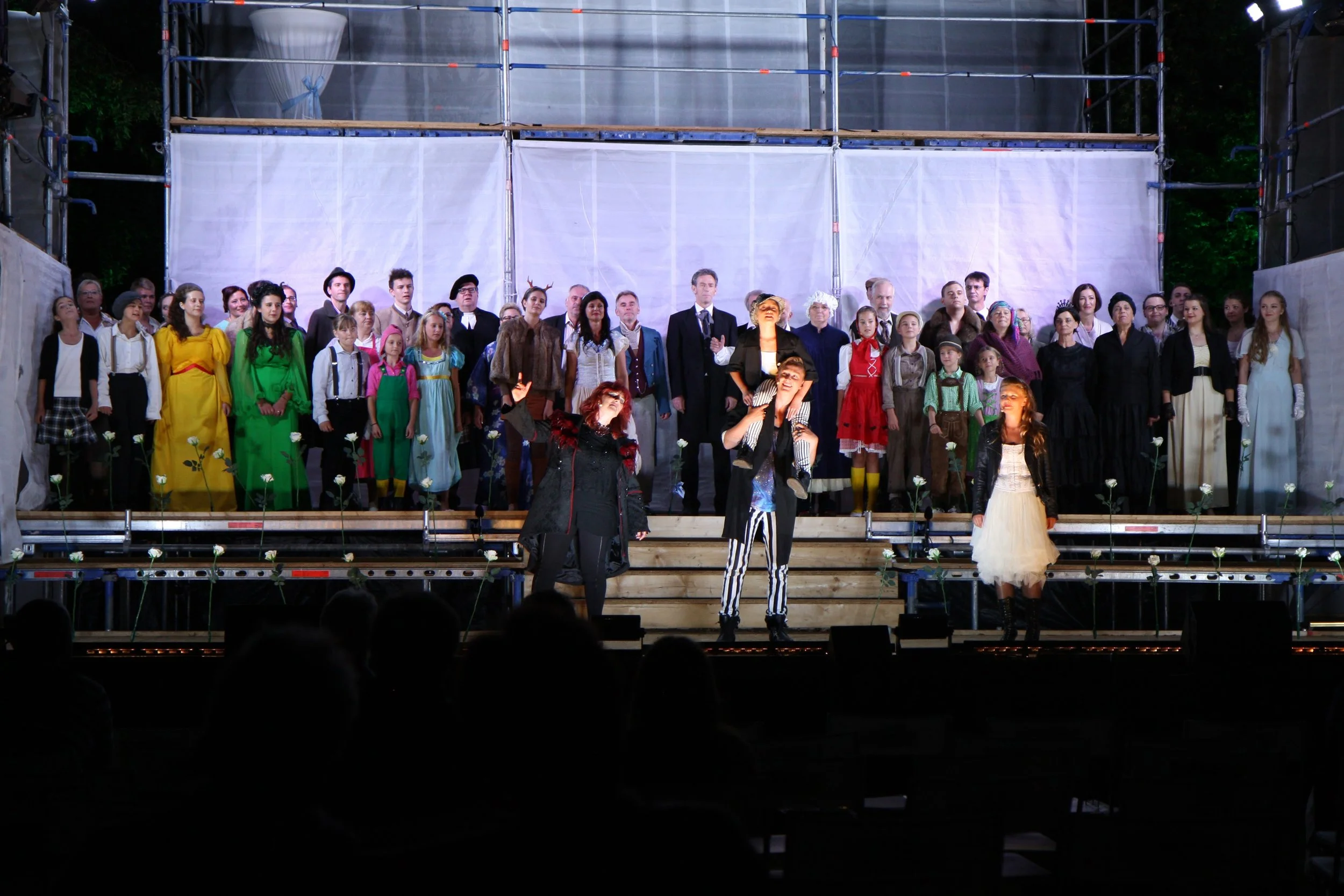 The image shows a theater stage with a large group of people performing in costumes, some dressed as fairy tale characters, while others are in formal or vintage clothing. The stage is decorated with flowers and the audience is visible in the foregro