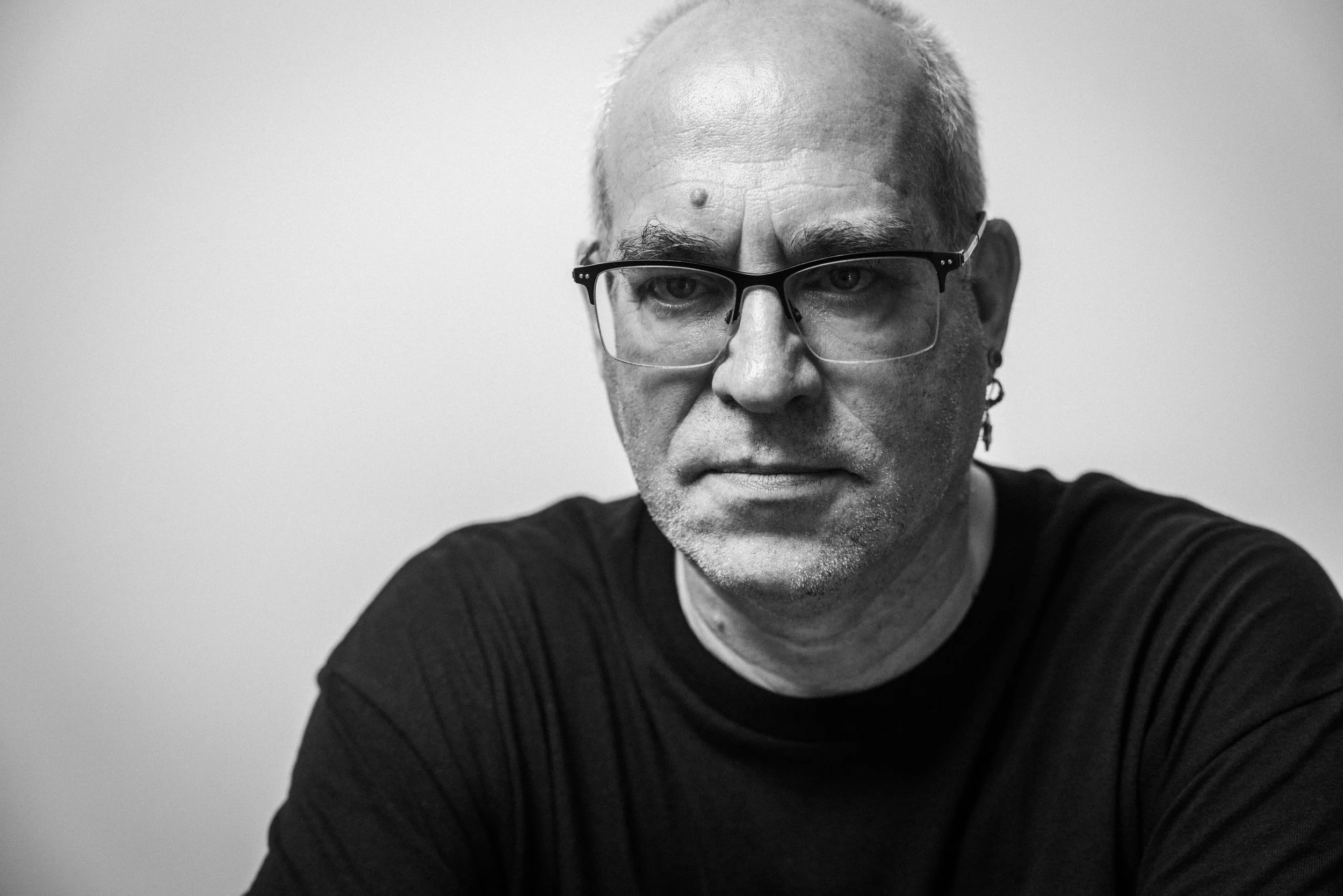 Schwarz-weiß-Fotografie eines Mannes mit Brille, Kurzhaar, skeptischem Ausdruck, trägt einen schwarzen Pullover.