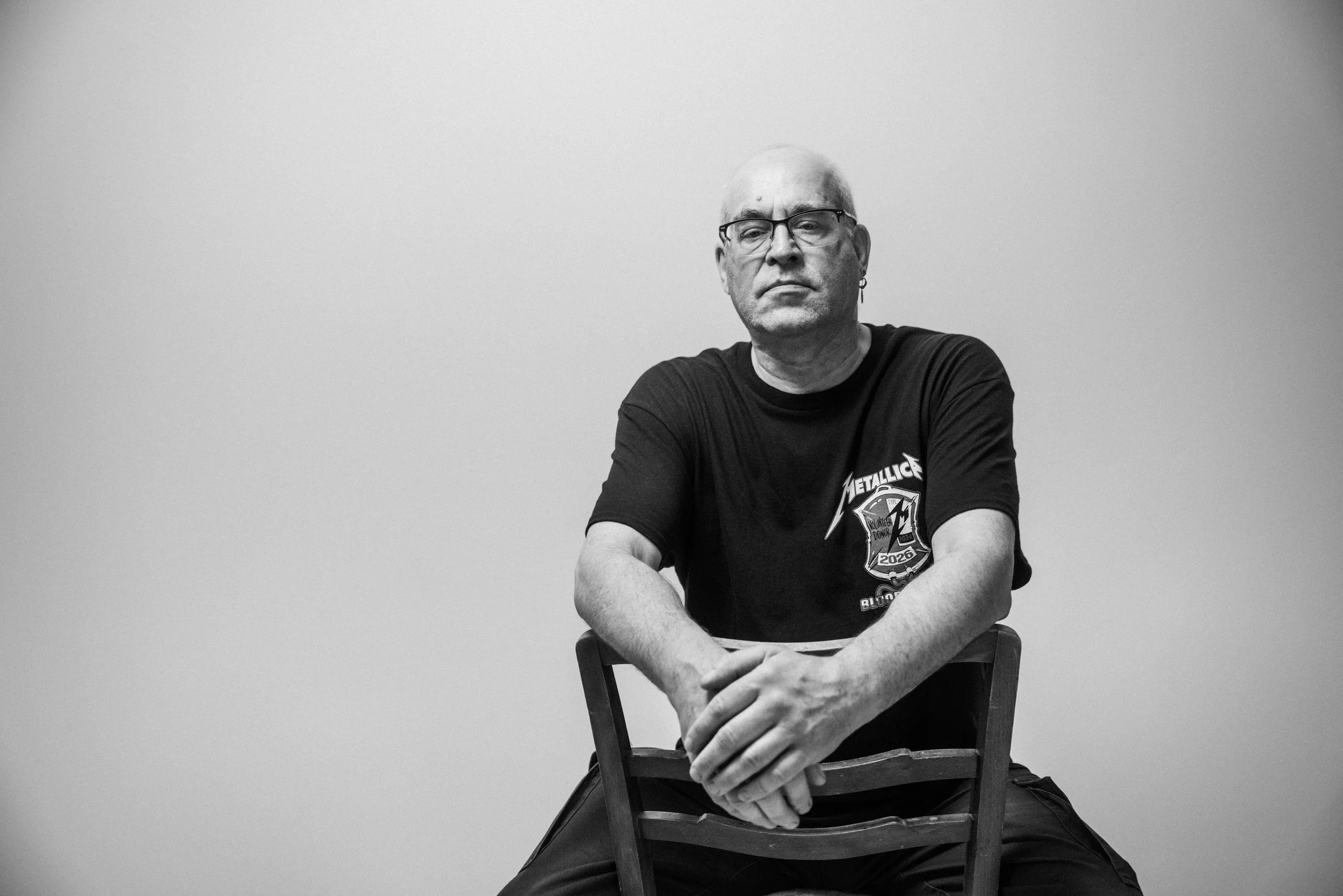 Ein älterer Mann mit Brille und Gliedern, trägt ein schwarzes T-Shirt mit einem Metallica-Logo, sitzt auf einem Stuhl vor einer hellen Wand, mit verschränkten Armen auf seinem Schoß.