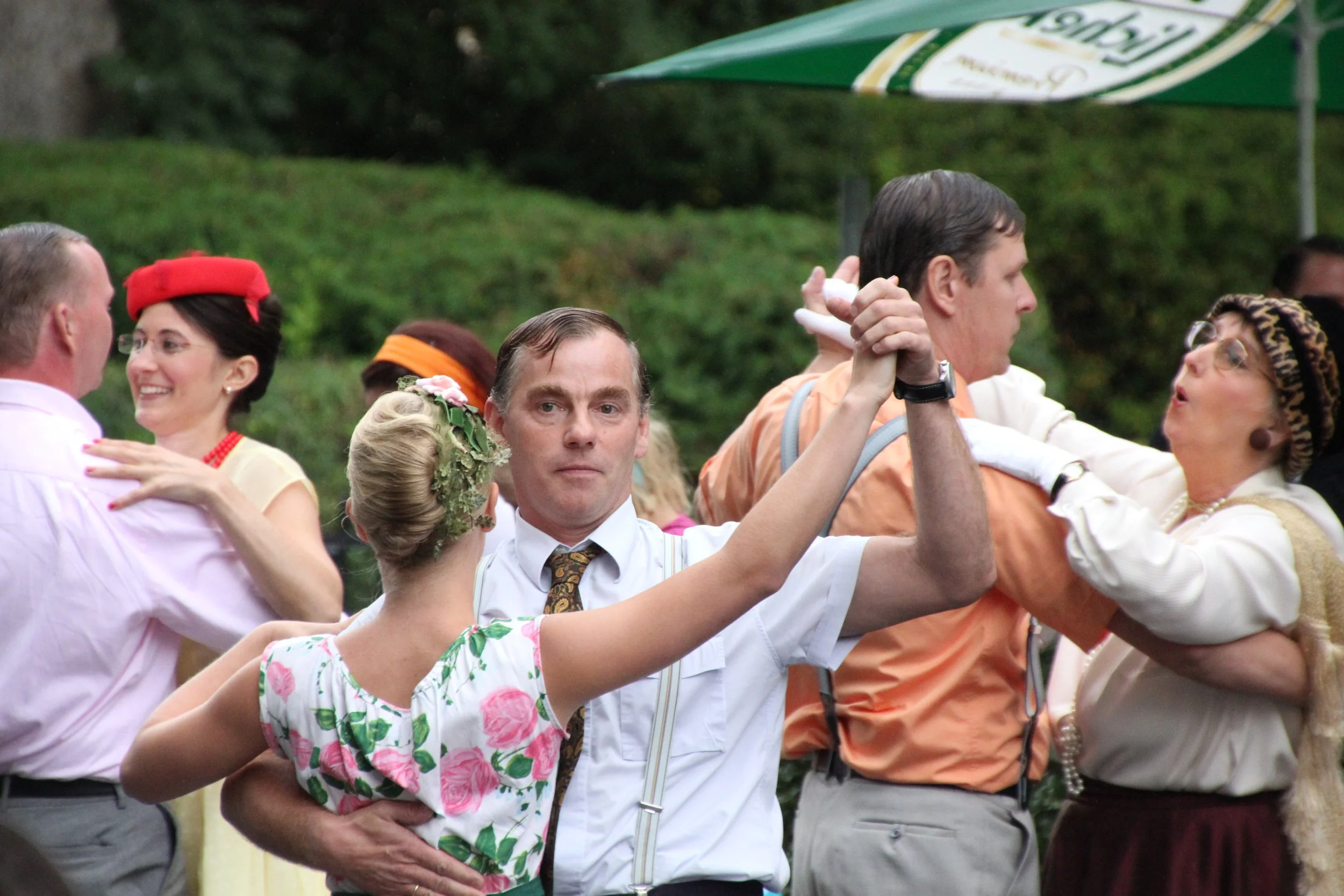Menschen tanzen im Freien, bekleidet in retro Kleidung, mit einem Mann im weißen Hemd im Vordergrund, der einen Tanzpartner hält, bei einer Frühlings- oder Sommerveranstaltung.