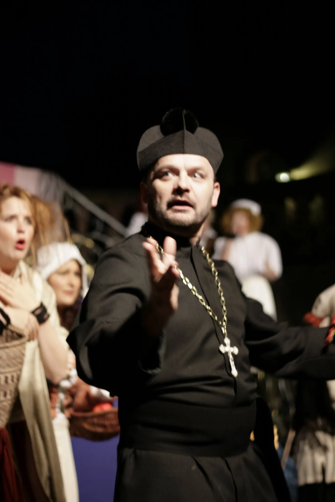 Ein Mann in schwarzer Kleidung mit einer großen goldenen Halskette und einem schwarzen Hut, der wie ein Priester oder religiöher Führer aussieht, steht im Mittelpunkt einer Menschenmenge, die verwirrt oder überrascht aussieht.