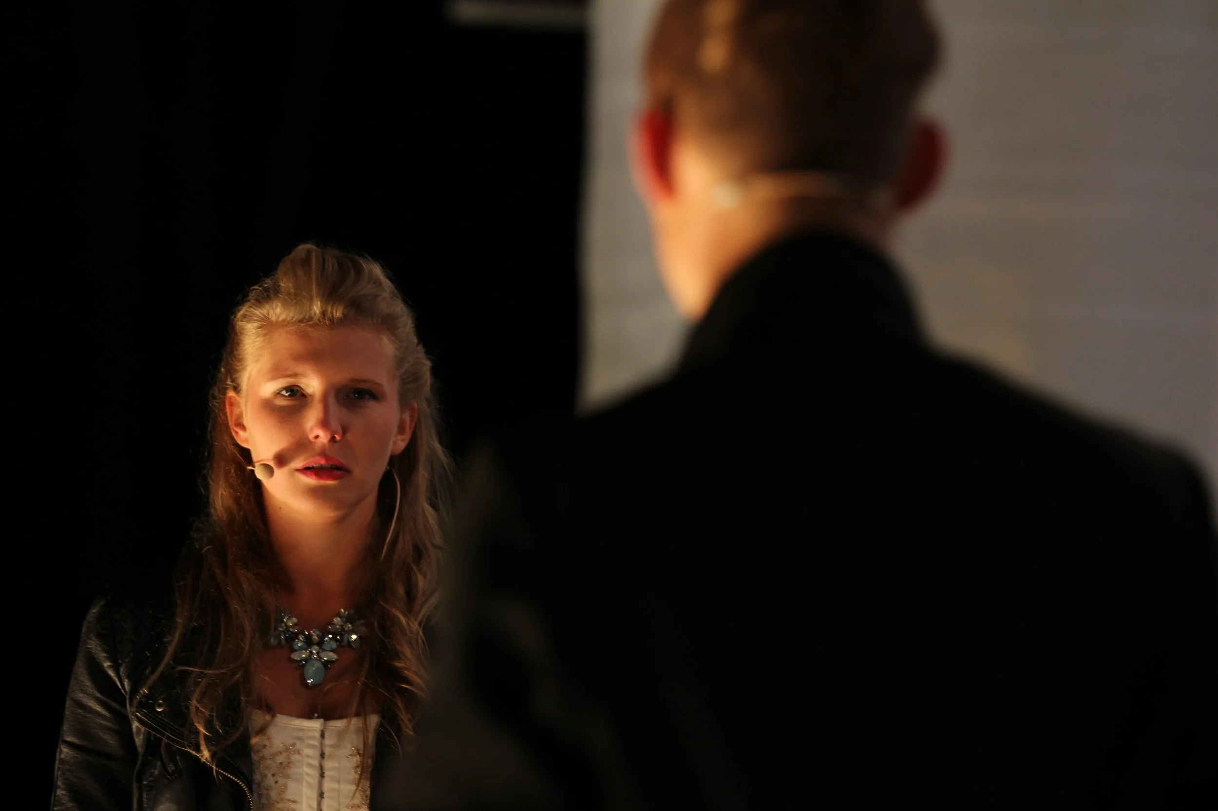 Eine junge Frau mit einem Headset und einer Halskette schaut nach vorne, während ein Mann mit dunklem Jackett im Vordergrund nur von hinten sichtbar ist. Das Ensemble der Birsteiner Festspiele 2016 und 2017 in: Der Wilde Grimm - Das Musical