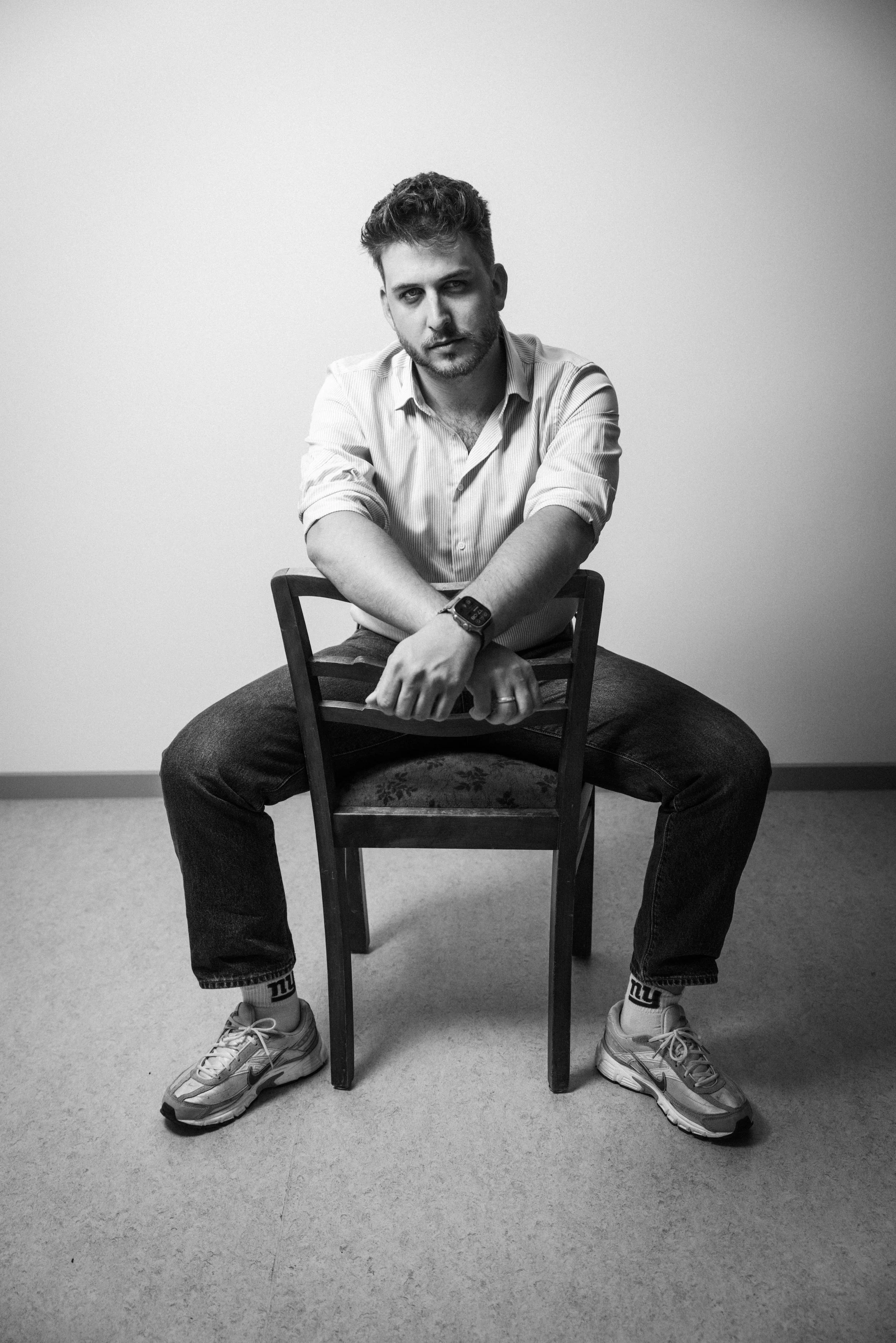 Schwarz-weiß Foto eines jungen Mannes, der auf einem Stuhl sitzt, mit verschränkten Armen und Blick zur Kamera, in einem schlichten Raum. Intendant Leon Roskoni in  in  Das Wilde Weib – Das Musical · Birsteiner Festspiele 2027