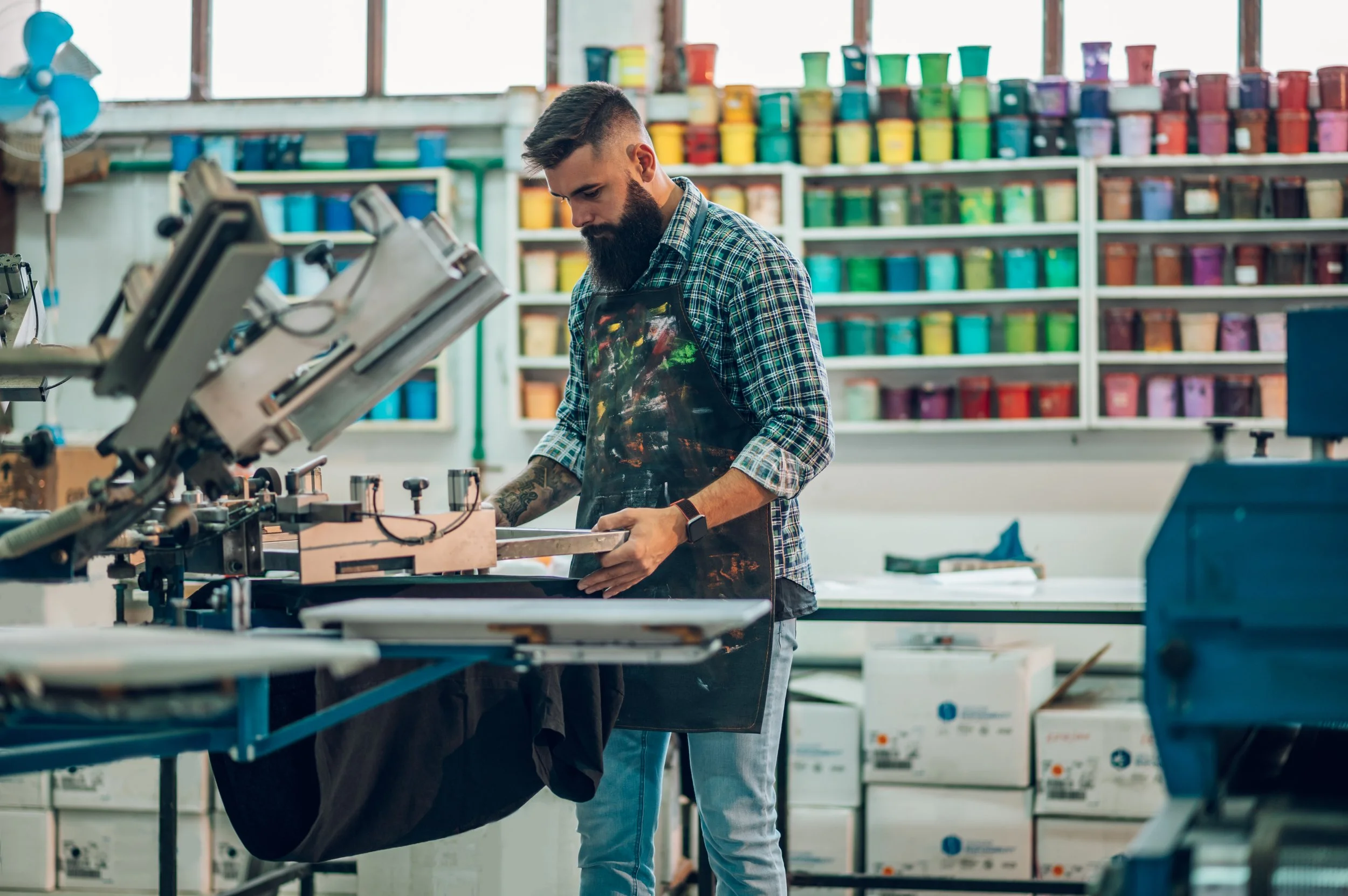 Ein Mann arbeitet in einer Werkstatt oder einem Kunststudio, umgeben von Farbbehältern auf Regalen im Hintergrund.
