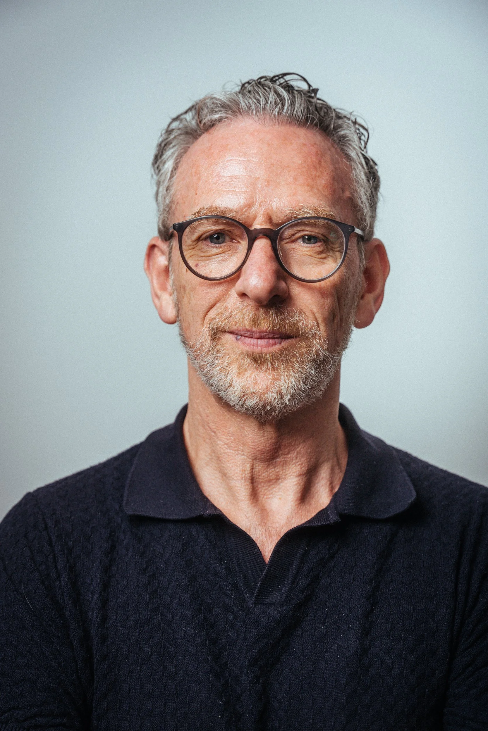 Ein älterer Mann mit grauem, lockigem Haar, Bart und Brille, trägt ein schwarzes, kabelgebundenes Oberteil, blickt leicht lächelnd in die Kamera vor einem hellen Hintergrund.