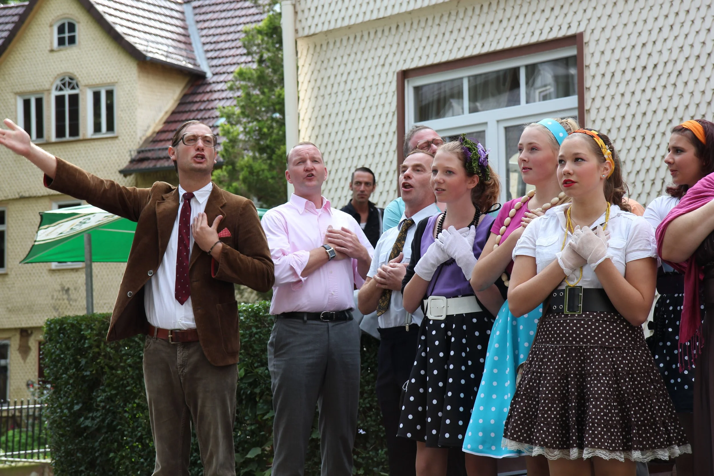 Eine Gruppe von Menschen, die im Freien singen und performen, einige mit Hand auf der Brust, in retro-inspirierter Kleidung, vor Häusern mit Gärten im Hintergrund.