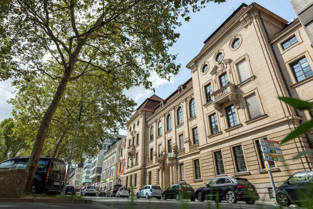 Historisches Gebäude mit mehreren Stockwerken und klassischen architektonischen Details, parkende Autos und ein Baum im Vordergrund auf einer urbanen Straße.