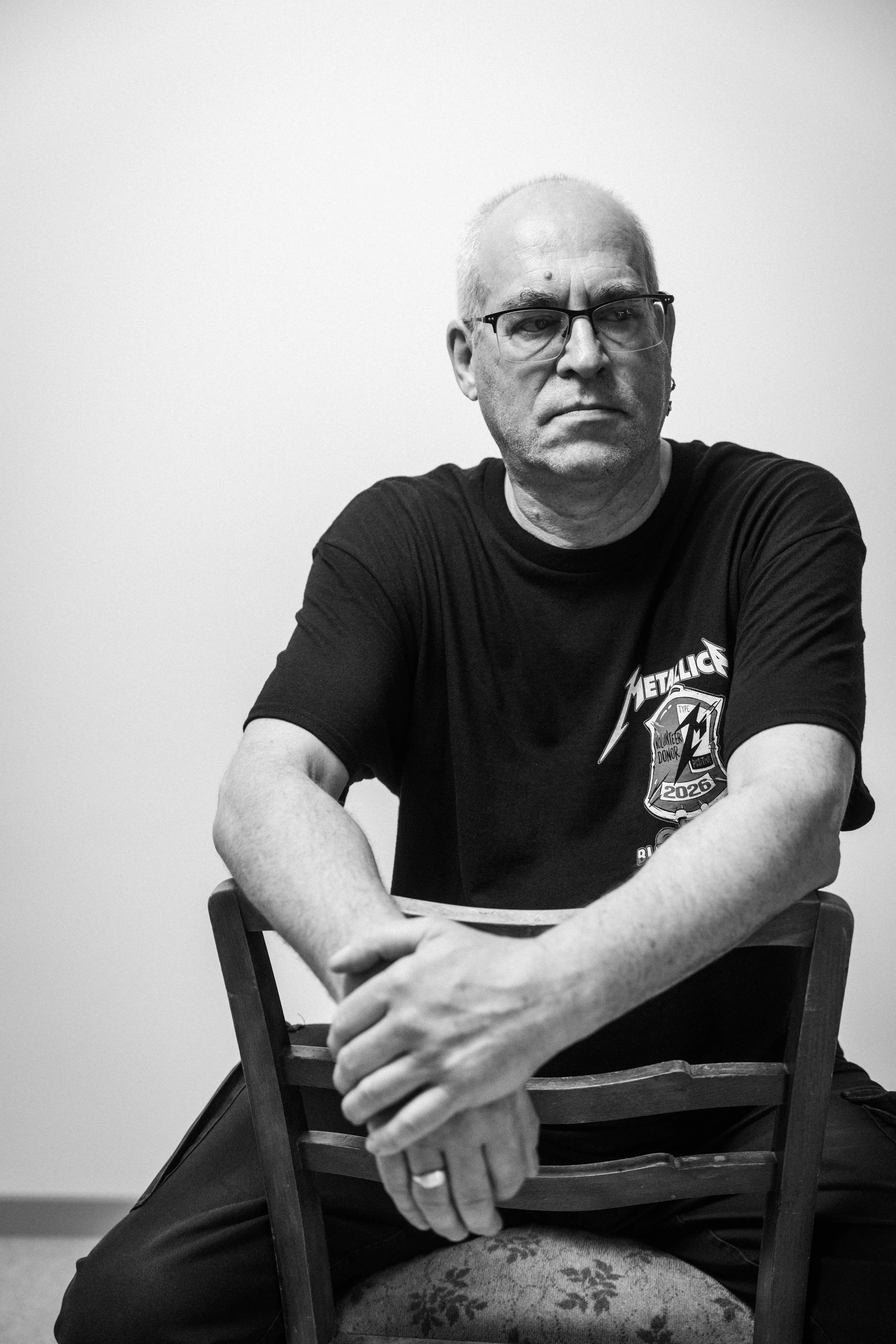 Schwarz-Weiß-Foto eines Mannes mit Brille, gepierctem Ohr und grauem Haar, der auf einem Stuhl sitzt. Er trägt ein schwarzes T-Shirt mit einem Metallica-Logo und einem Shield mit der Aufschrift 2026. Der Mann schaut nach rechts und wirkt ernst.