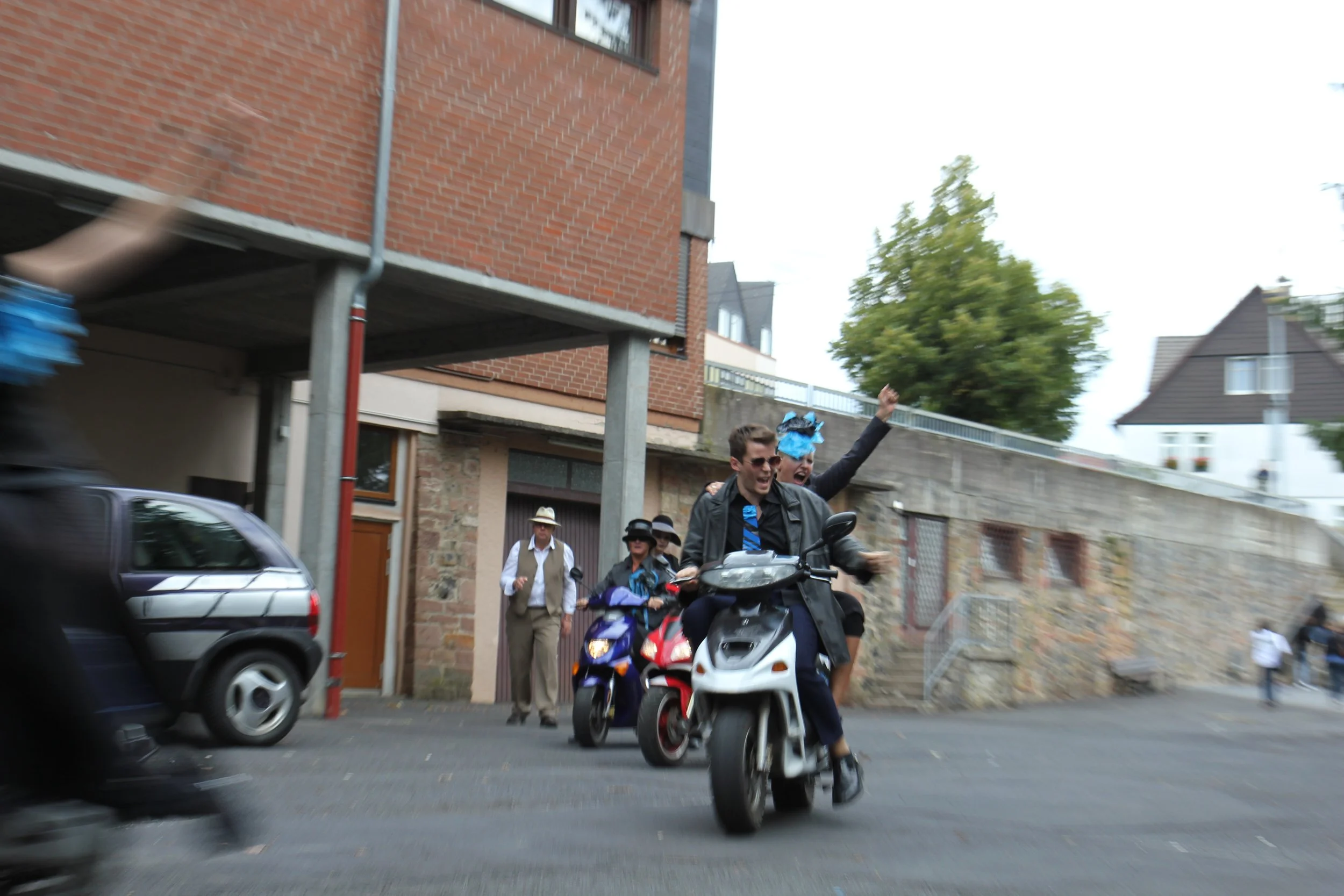 Zwei Männer fahren lachend auf einem weißen Scooter, einer trägt eine blaue Perücke und beide jubeln. Im Hintergrund stehen mehrere Menschen, einige in Hüten, vor einem Haus mit Stein- und Ziegelmauer.