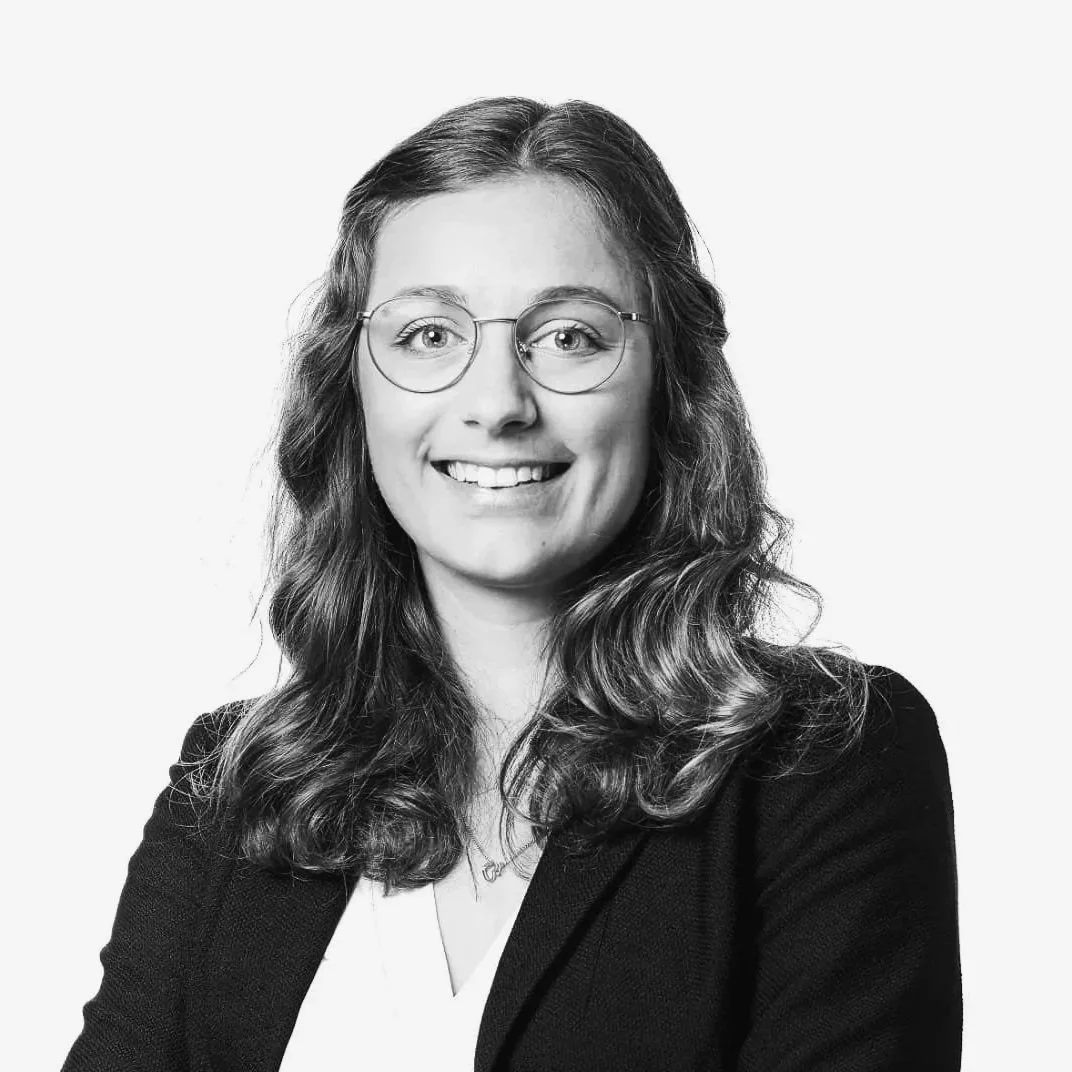 Schwarz-Weiß-Porträt einer lachenden Frau mit Brille, lockiger langer Haar, schwarzer Jacke und weißer Bluse, vor hellem Hintergrund.