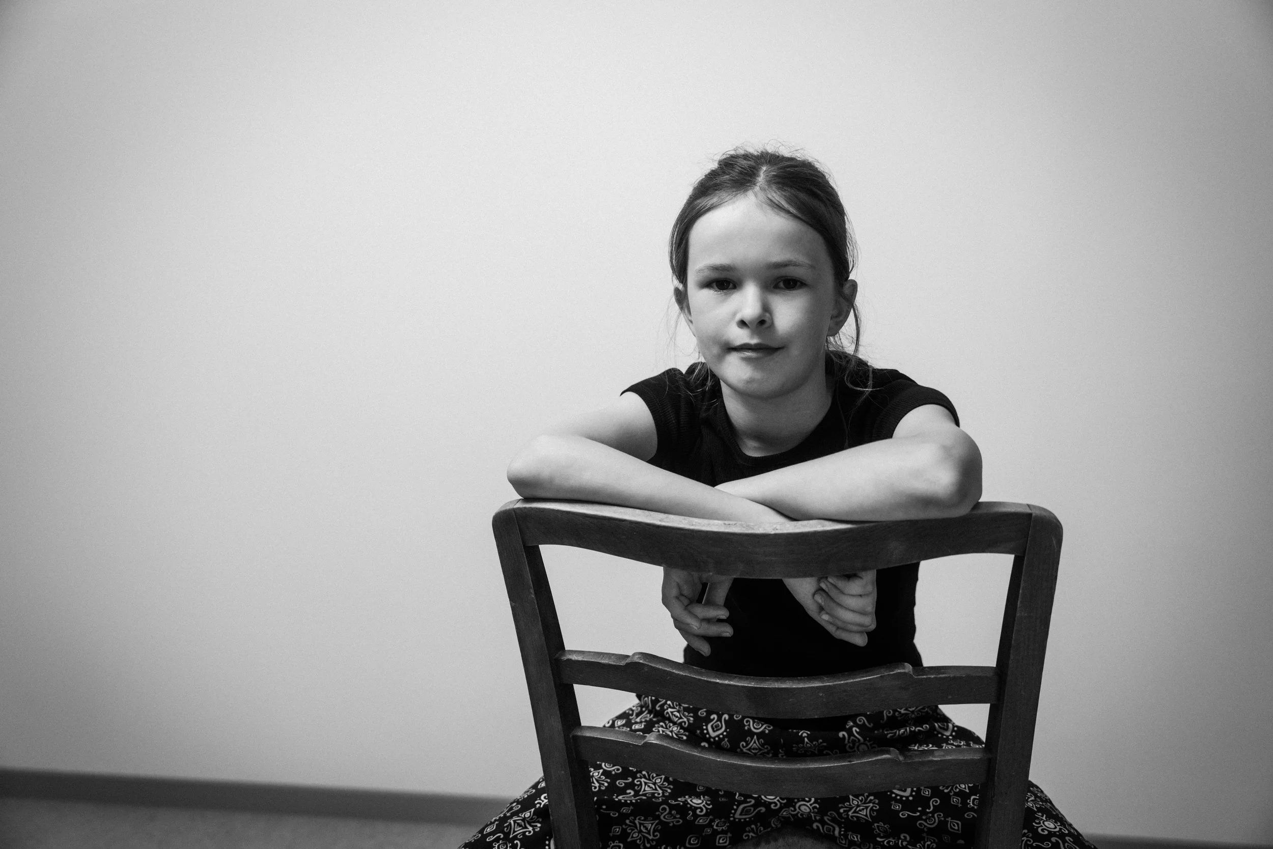 Schwarz-Weiß-Foto eines jungen Mädchens, das auf einem Stuhl sitzt und die Arme auf die Rückenlehne legt, vor einer leeren Wand.