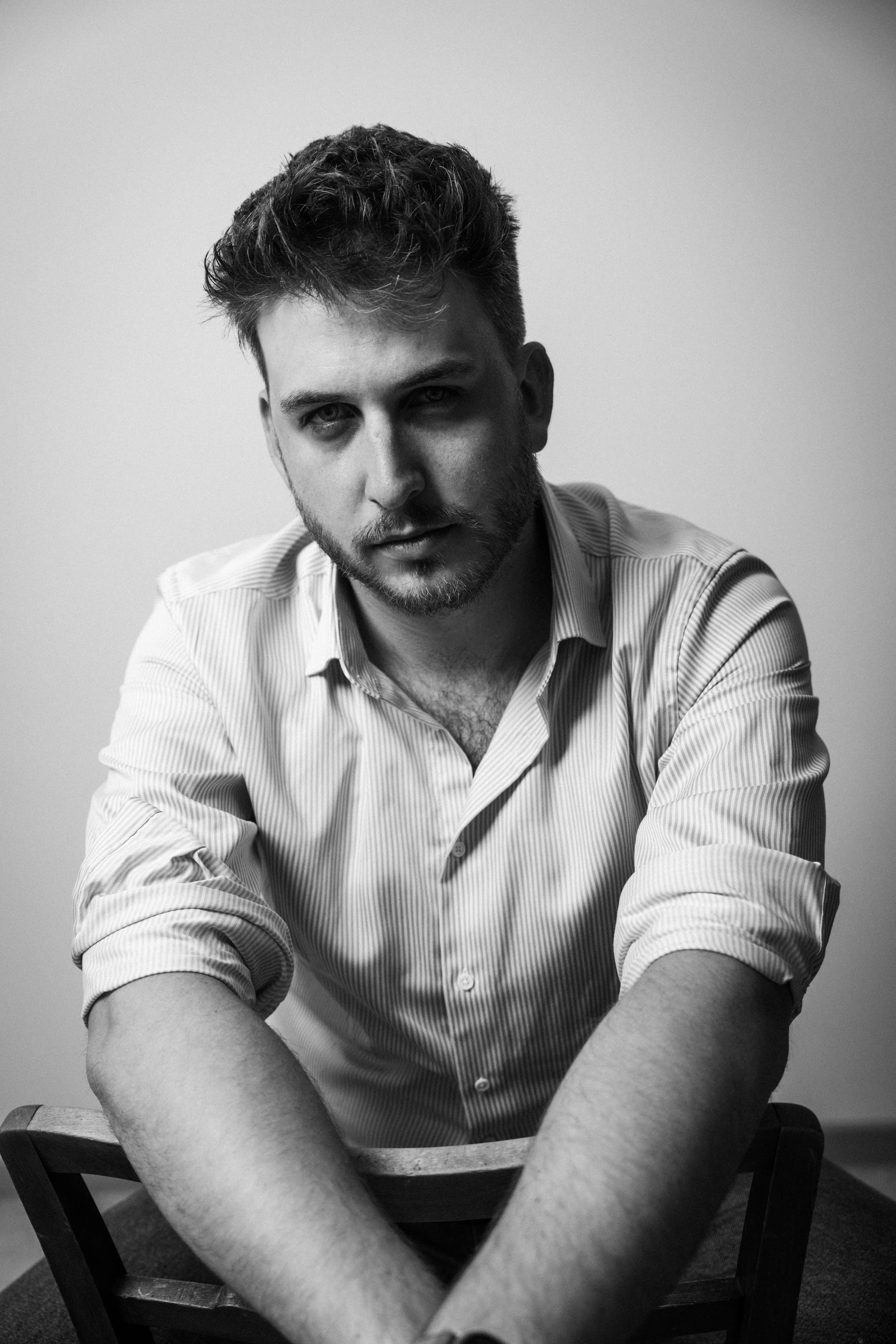 Schwarz-Weiß-Foto eines jungen Mannes, der auf einem Stuhl sitzt, mit einem ernsten Gesichtsausdruck, trägt ein Hemd mit Streifenmuster, seine Arme sind auf dem Tisch vor ihm. Der Hintergrund ist schlicht.
