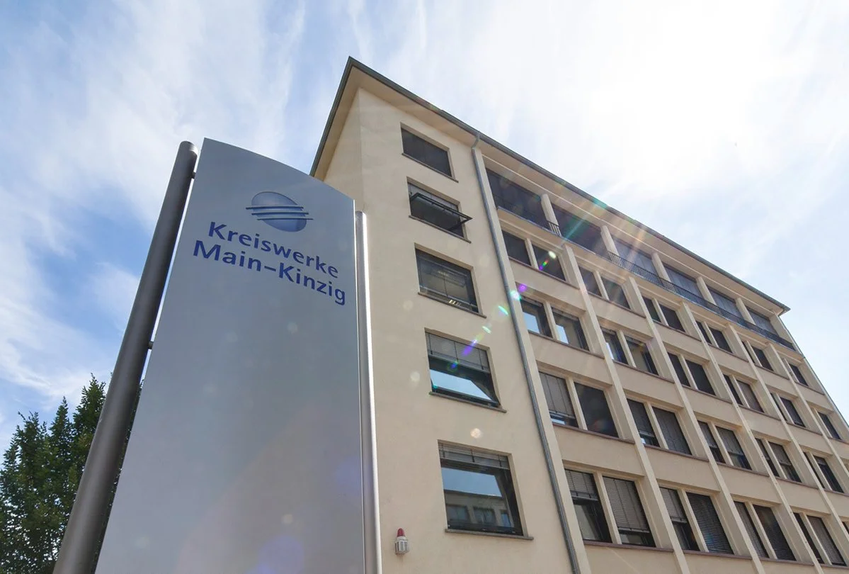 Ein mehrstöckiges Gebäude mit einem weißen Fassaden und mehreren fenstern, daneben ein Schild mit dem Logo und dem Namen 'Kreiswerke Main-Kinzig'.