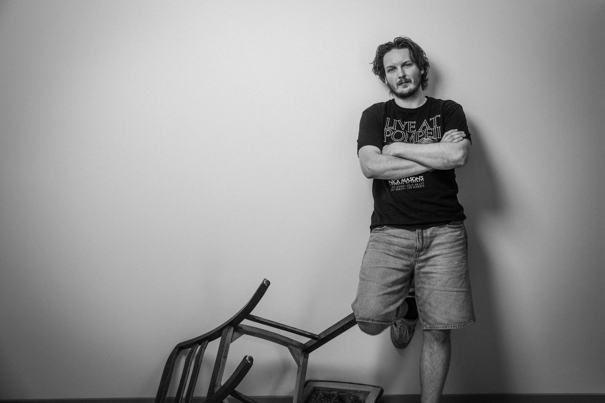 Schwarz-weiß Foto eines Mannes mit lockigem Haar und Bart, der gegen eine weiße Wand lehnt, Arme verschränkt trägt ein T-Shirt mit der Aufschrift »Live at Pompeii« und Shorts, neben ihm liegt ein umgestürzter Stuhl.
