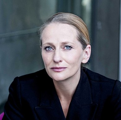 Porträt einer ernst aussehenden Frau mit blonden Haaren und blauen Augen, die einen schwarzen Blazer trägt, vor einer glasschweren, grünen Wand.
