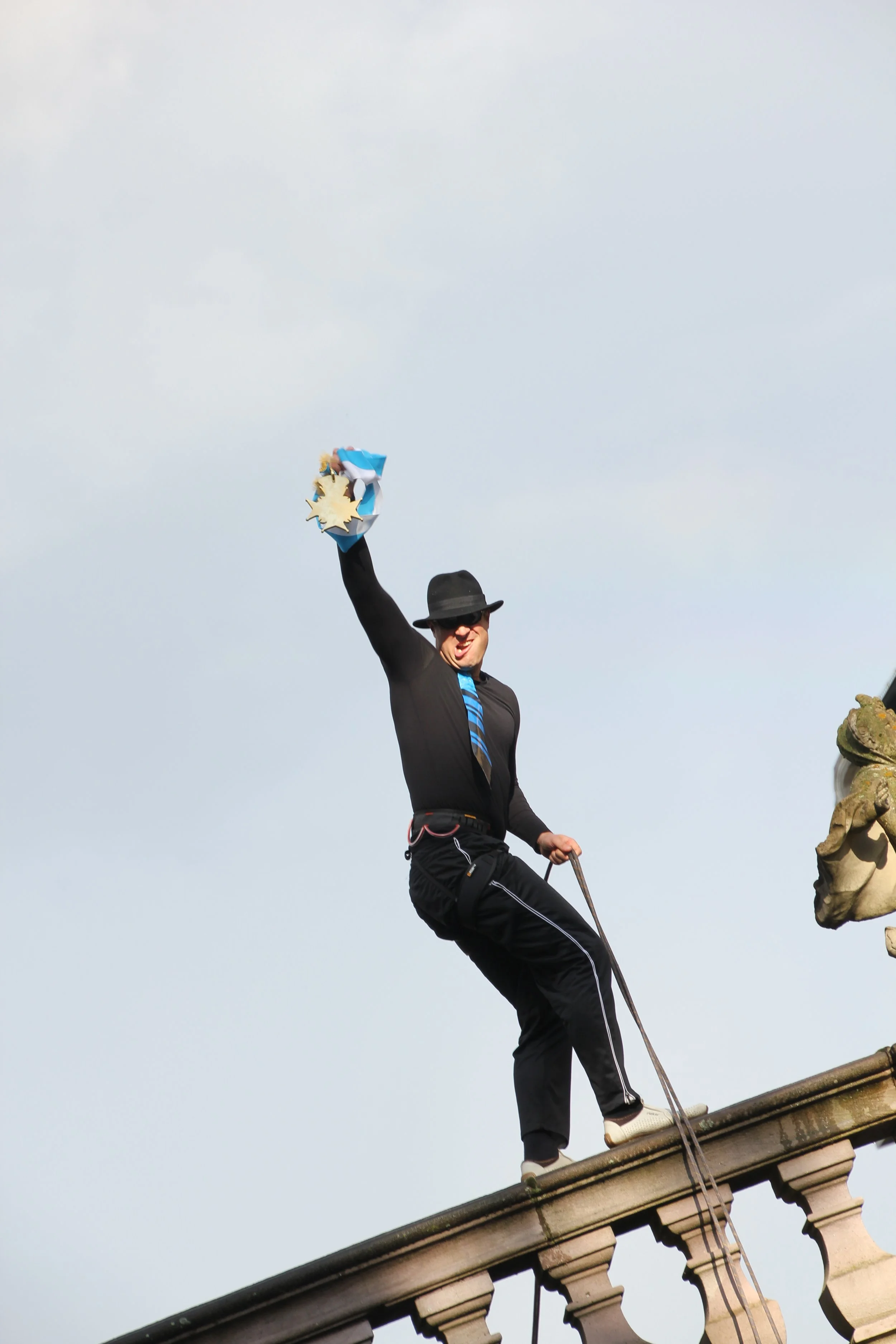Ein Mann in schwarz mit Hut, Krawatte und Sonnenbrille steht auf einer Dachkante, hält eine Packung mit goldenen Sternen und blauer Verpackung hoch und freut sich.