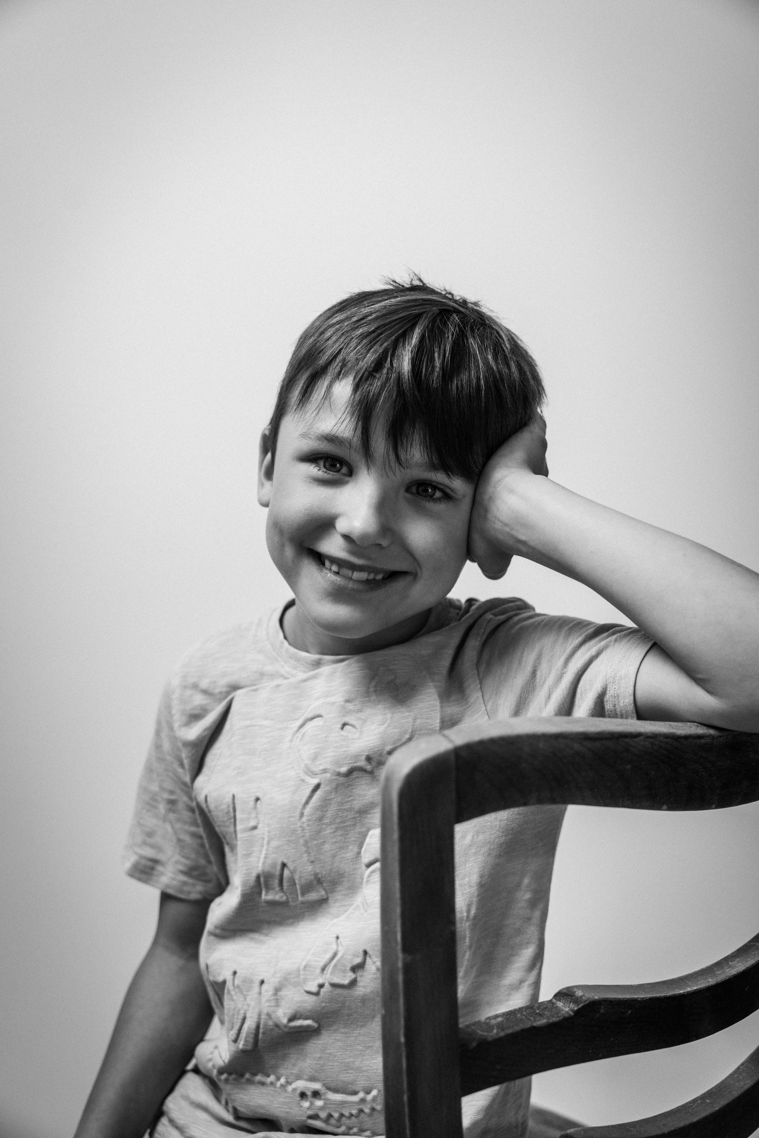 Schwarz-weiß-Foto eines lachenden Jungen mit kurzen Haaren, der auf einem Stuhl sitzt und die Hand an den Kopf legt.