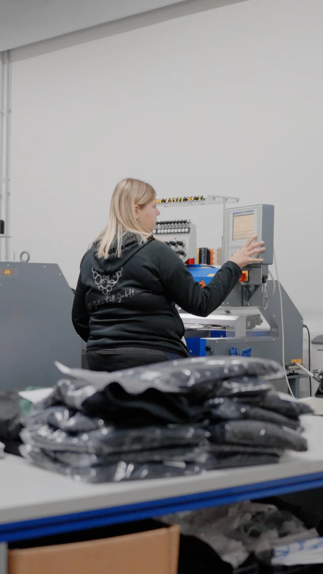Eine Frau arbeitet an einer Maschine in einer Werkstatt, im Vordergrund sind schwarze, verpackte Gegenstände auf einem Tisch.