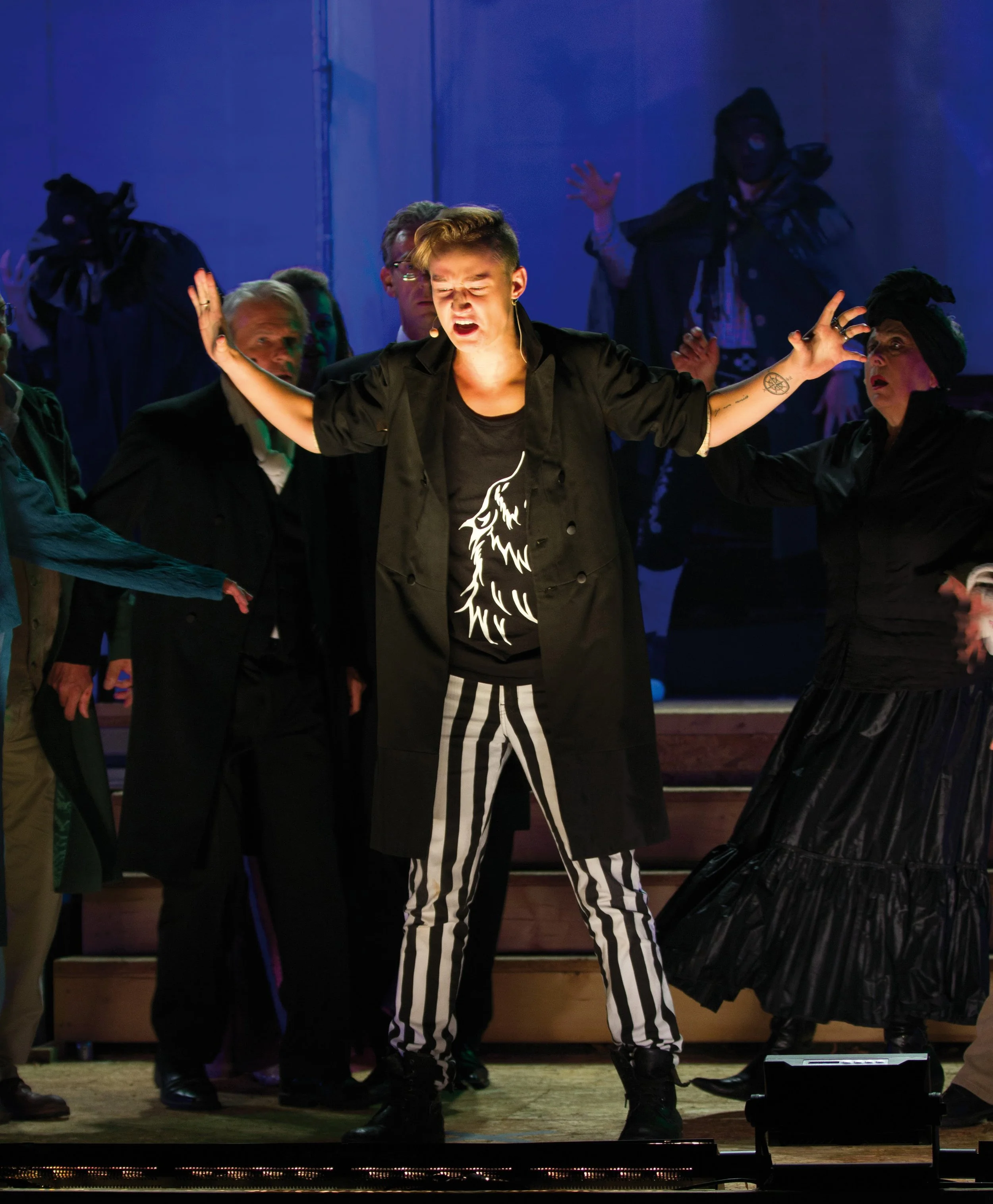 Schauspielerin bei einer Theateraufführung mit dramischem Ausdruck, umgeben von anderen Darstellern in dunkler Kleidung auf der Bühne.