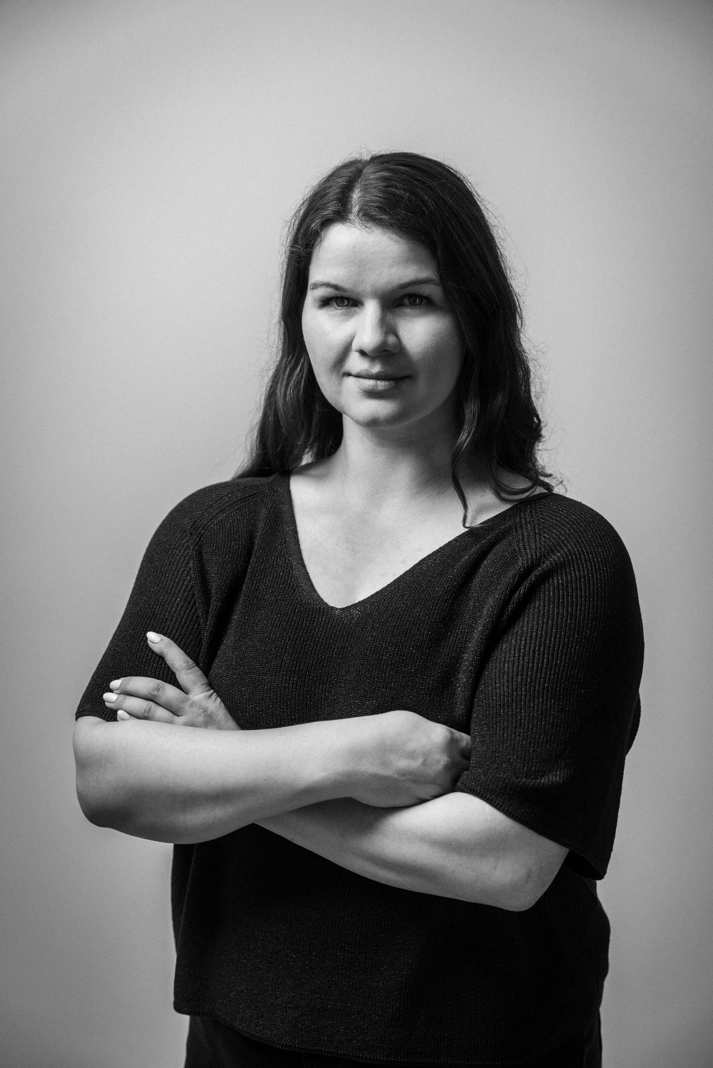 Schwarz-weiß-Foto einer Frau mit dunklen, mittellangen Haaren, die die Arme verschränkt trägt, vor einem grauen Hintergrund. Elina Beer in  in  Das Wilde Weib – Das Musical · Birsteiner Festspiele 2027