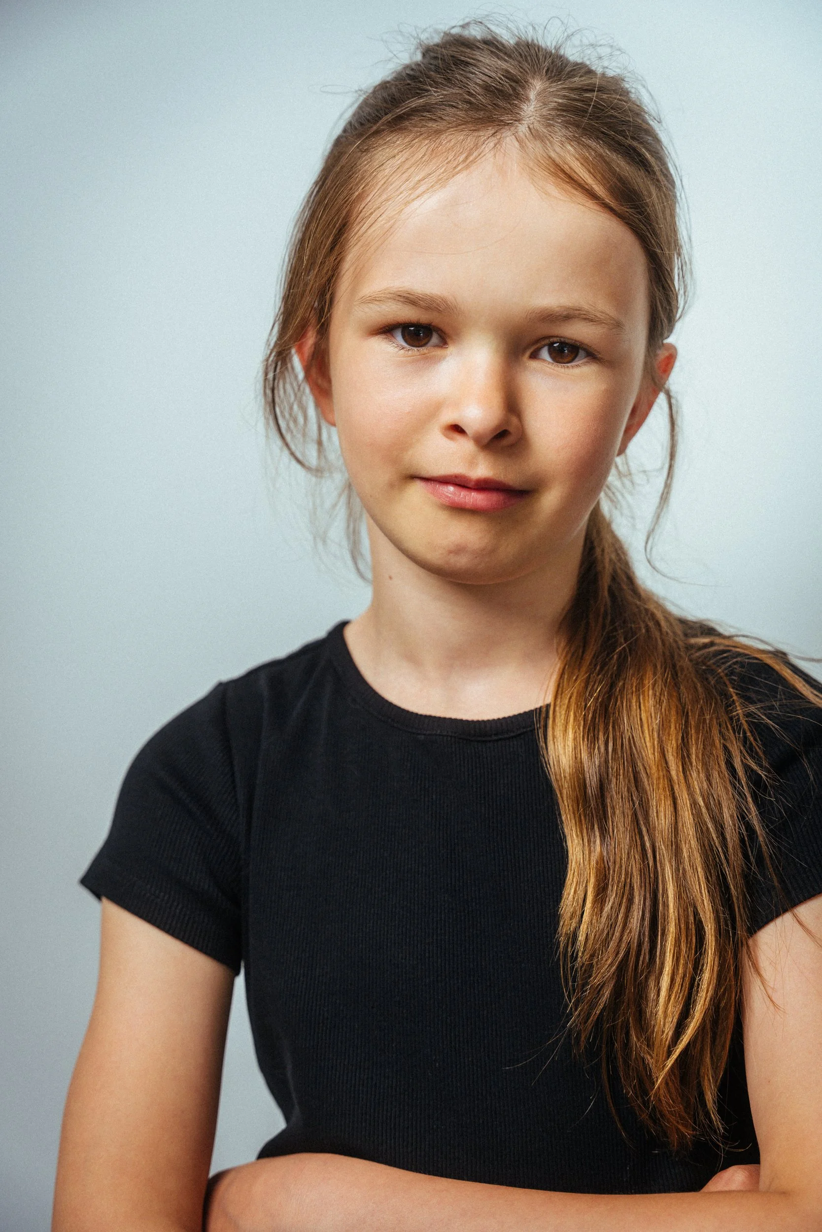 Ein junges Mädchen mit langen, hellbraunen Haaren, die nach rechts fallen, trägt ein schwarzes T-Shirt und hat einen neutralen Gesichtsausdruck. Der Hintergrund ist hellgrau.