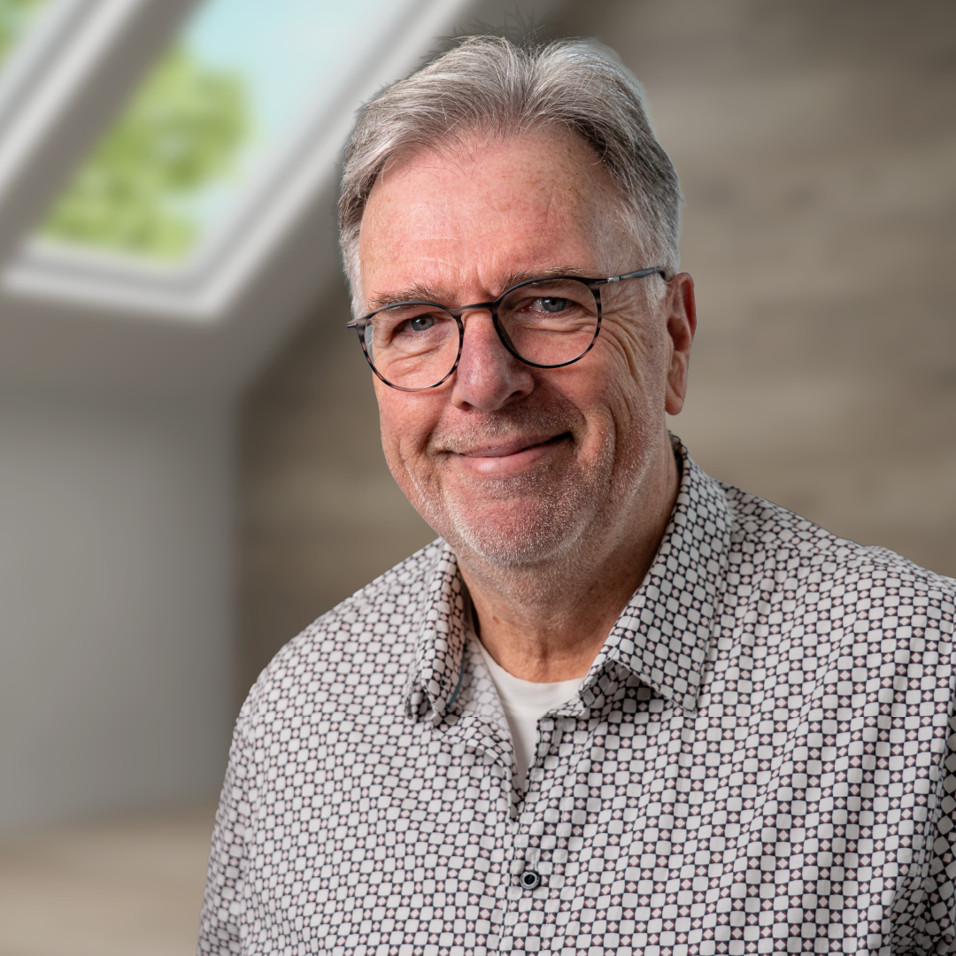 Ein freundlicher älterer Mann mit Brille, grauem Haar und einem karierten Hemd lächelt in die Kamera. Im Hintergrund ist ein Dachfenster und eine helle Wand.