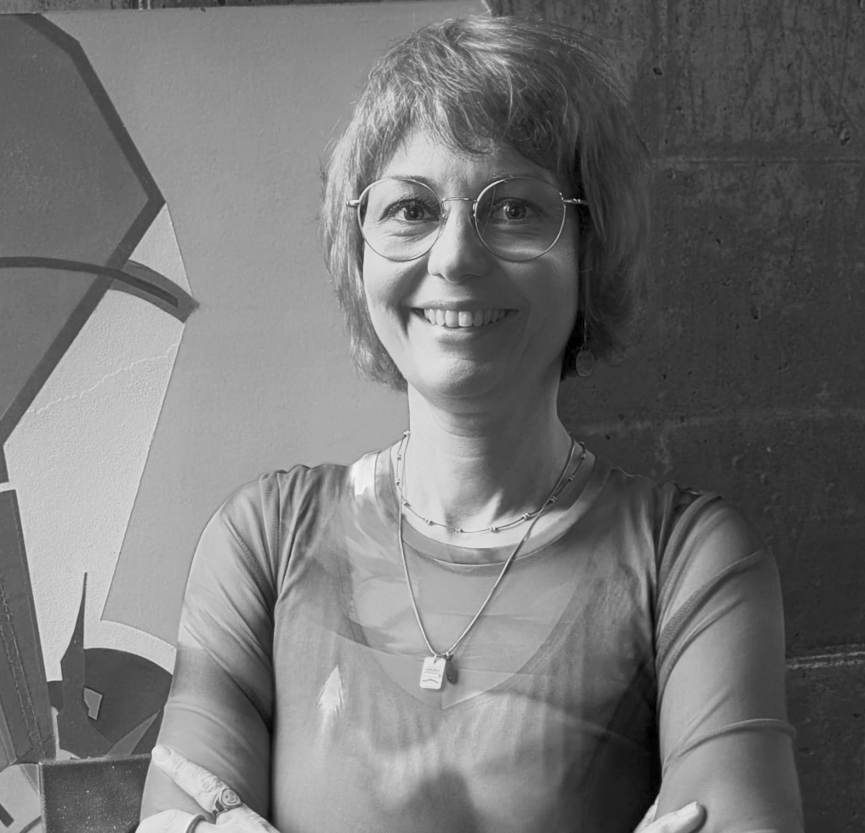 Eine lachende Frau mit kurzen, lockigen Haaren und Brille, trägt eine Kette und ein Top, steht vor einer Wand mit graffitiähnlicher Kunst. Schwarzweißbild.