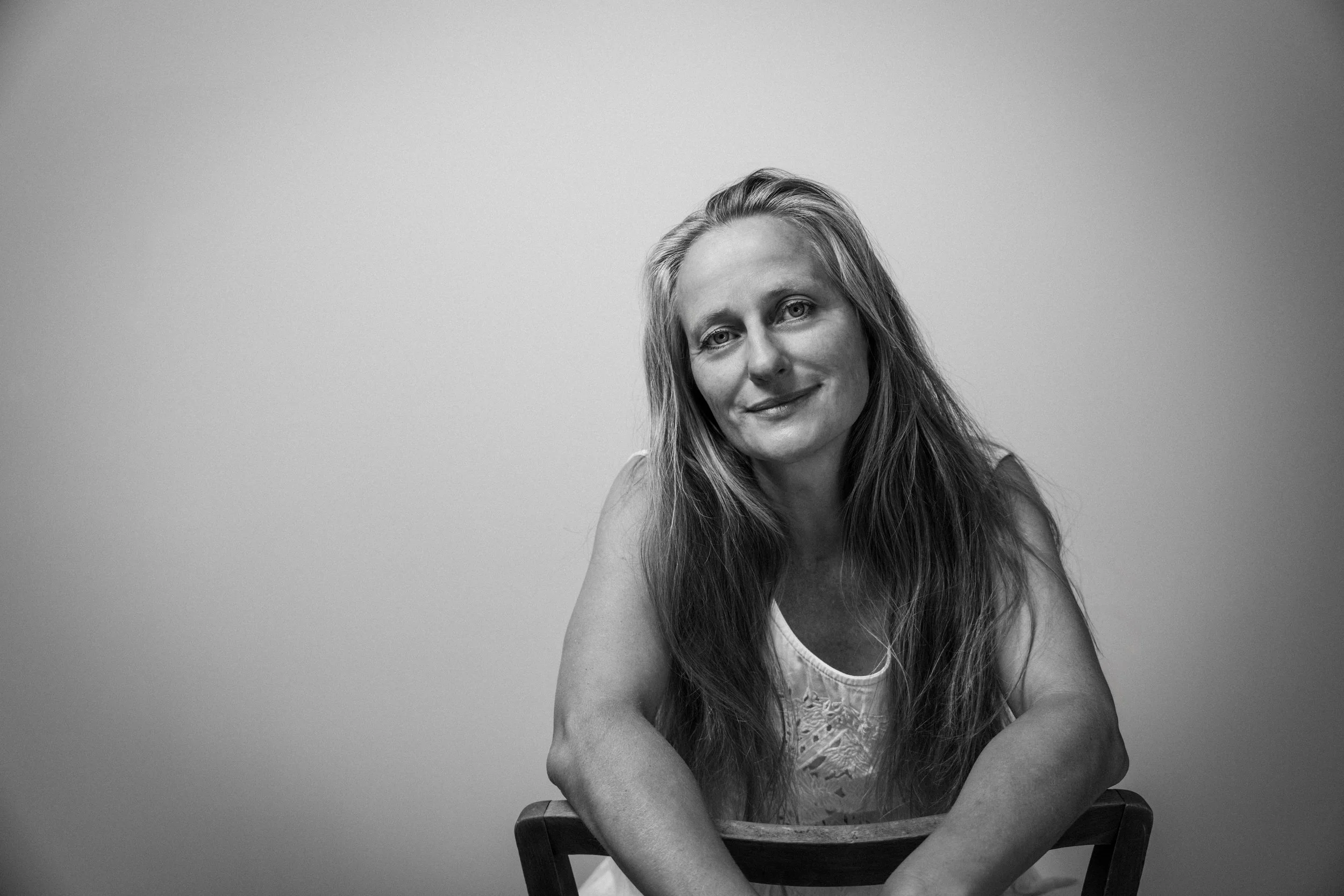 Regina Leitner, Regisseurin der Birsteiner Festspiele. Schwarz-weiß Porträt einer lächelnden Frau mit langen Haaren, sitzend auf einem Stuhl, vor einem hellgrauen Hintergrund.