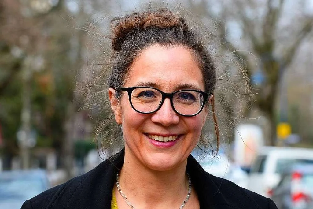 Eine lächelnde Frau mit Brille, dunklem Haar und informeller Kleidung im Freien, vermutlich in einer städtischen Umgebung, mit Bäumen und Autos im Hintergrund.