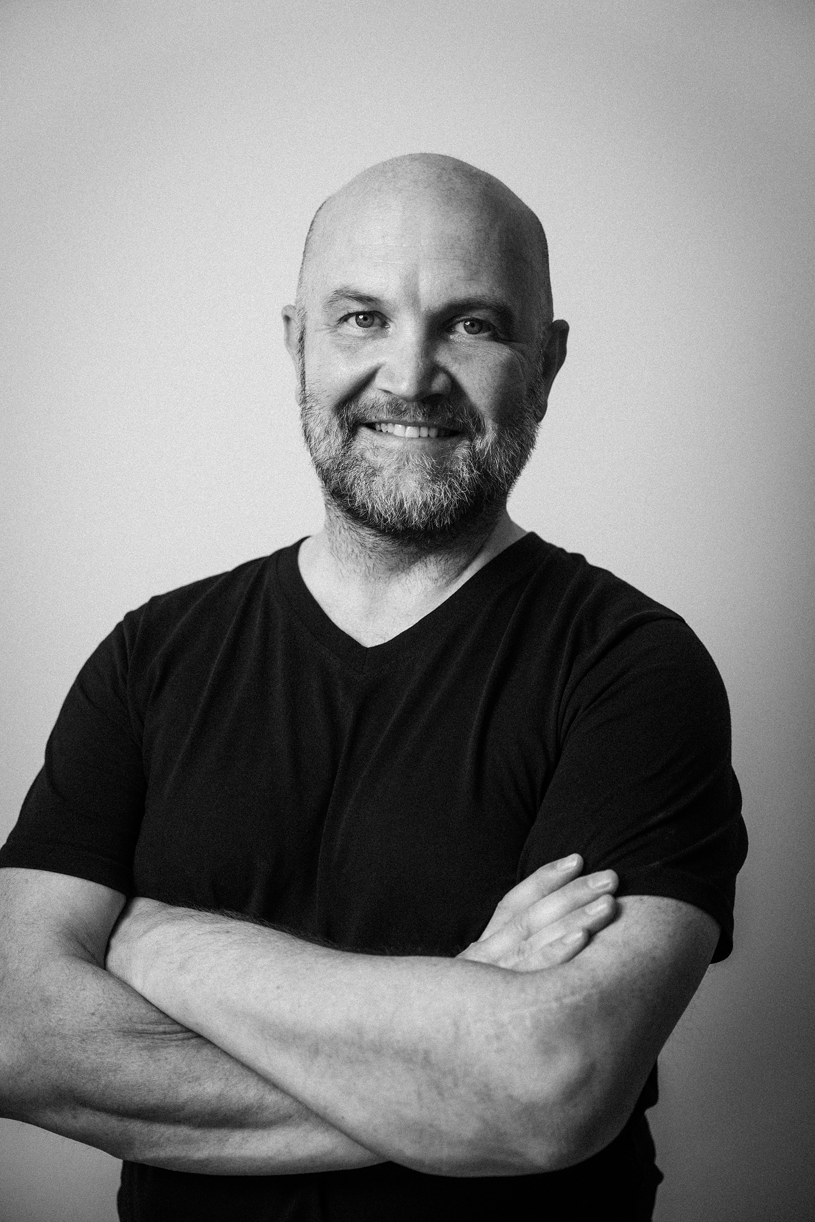 Schwarz-Weiß-Bild eines Mannes mit kurzen Haaren und Vollbart, lächelnd, trägt ein dunkles T-Shirt, Blick in die Kamera, Hintergrund grau, Arme verschränkt.