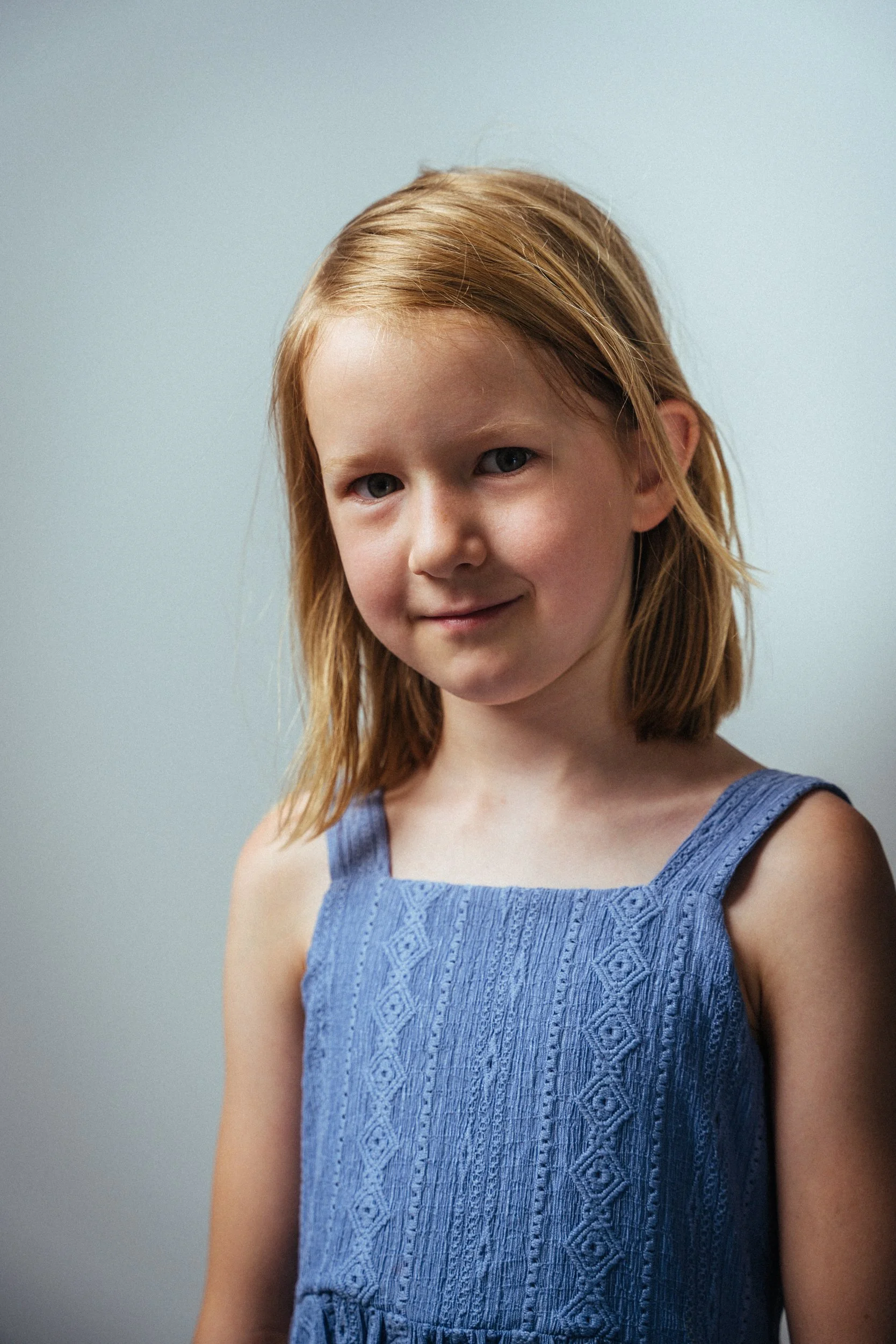 Ein junges Mädchen mit rotbraunen Haaren, das ein blaues ärmelloses Kleid trägt. Sie steht vor einem grauen Hintergrund und lächelt leicht in die Kamera.
