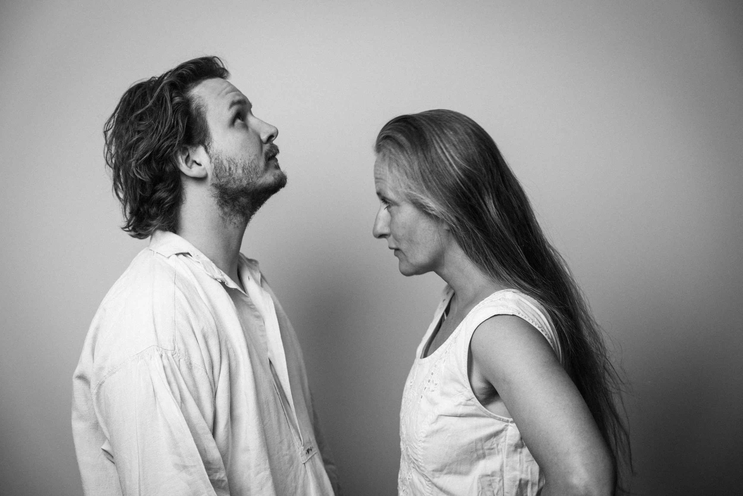 Schwarz-Weiß-Foto eines Mannes und einer Frau, die sich gegenüberstehen, mit nach oben gerichteten Blicken. Der Mann trägt ein Hemd, die Frau ein ärmelloses Oberteil. Sie sind nahe beieinander, auf grauem Hintergrund.