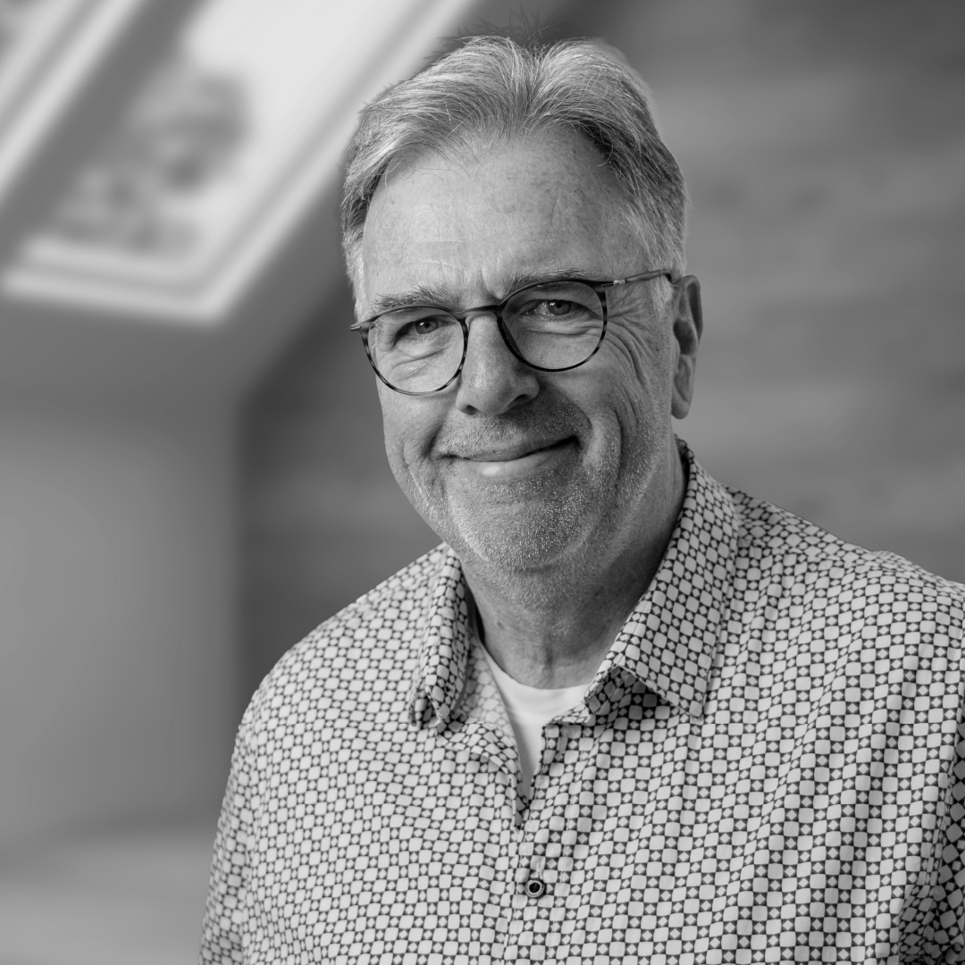 Ein lächelnder älterer Mann mit Brille, grauem Haar und Bart trägt ein geometrisch gemustertes Hemd. Im Hintergrund ist ein Bildschirm sichtbar.