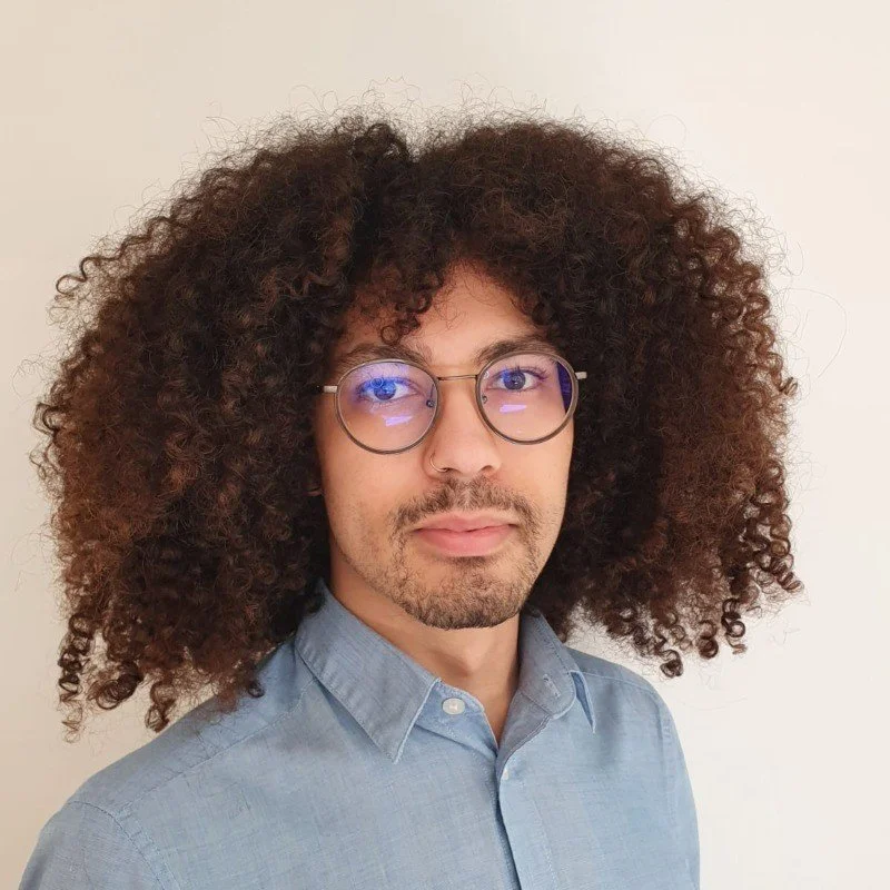 Junge Mann mit runden Brillen, lockigem Bart und großen lockigen Haaren trägt ein hellblaues Hemd vor einem neutralen Hintergrund.