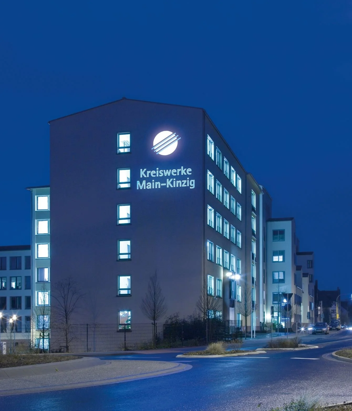 Ein mehrstöckiges Gebäude bei Nacht mit im Licht erleuchteten Fenstern und einem beleuchteten Firmenlogo an der Fassade, das die Kreisewerke Main-Kinzig zeigt.